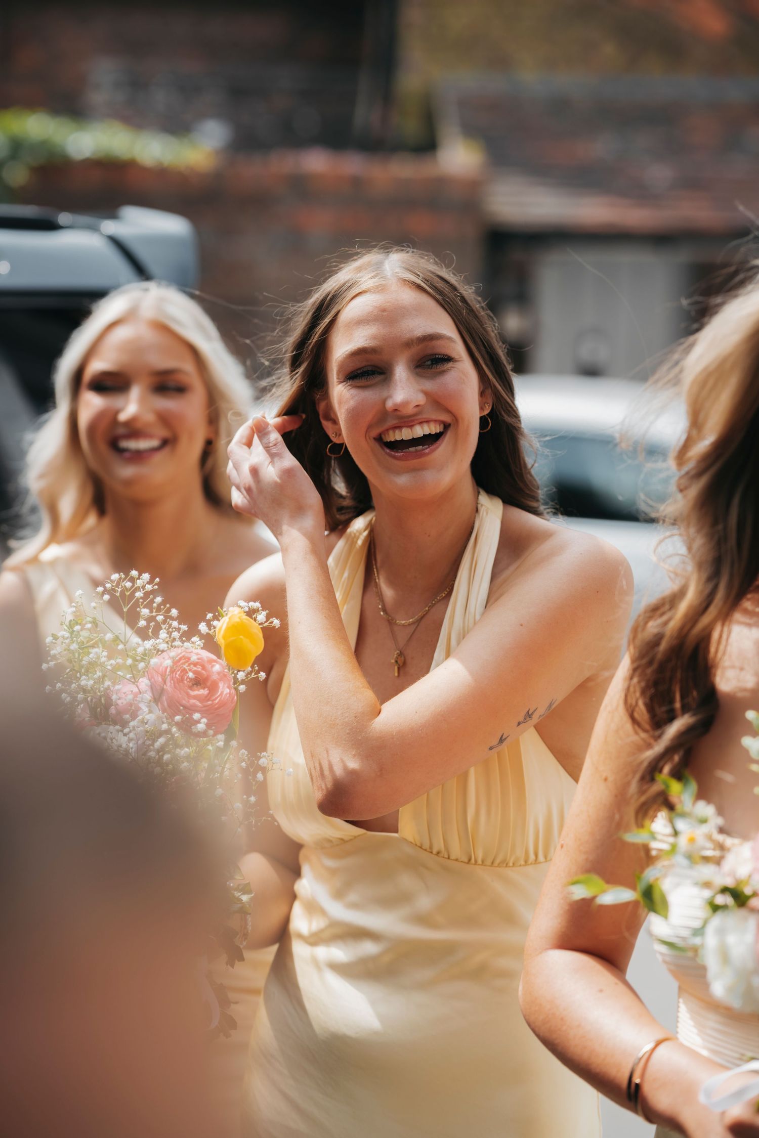 Laughing bridesmaid in a pale yellow satin dress tucks her hair back while smiling friends hold bouquets nearby.