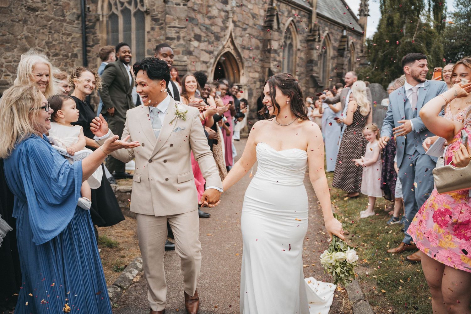 Newlyweds hold hands and walk down a church path as joyful guests cheer and applaud around them.