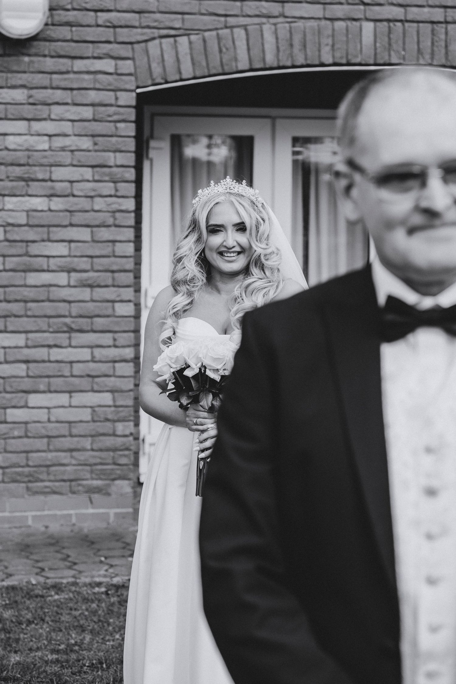 Black and white photo of a smiling bride in a tiara and veil watching her father from behind a doorway.
