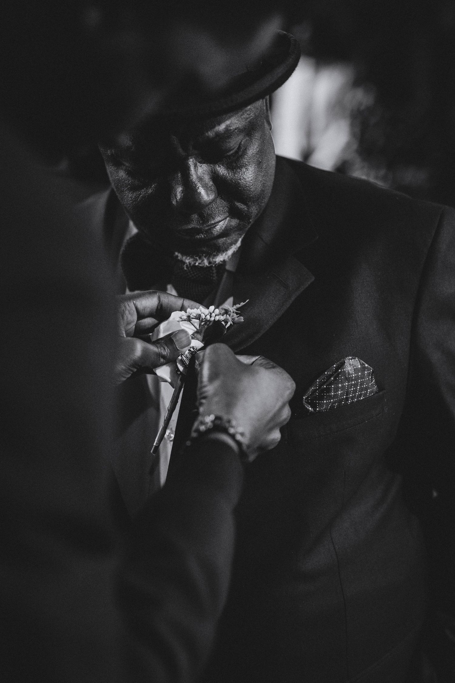 Black and white portrait of a man in a hat and suit having a boutonniere pinned to his lapel before a wedding.