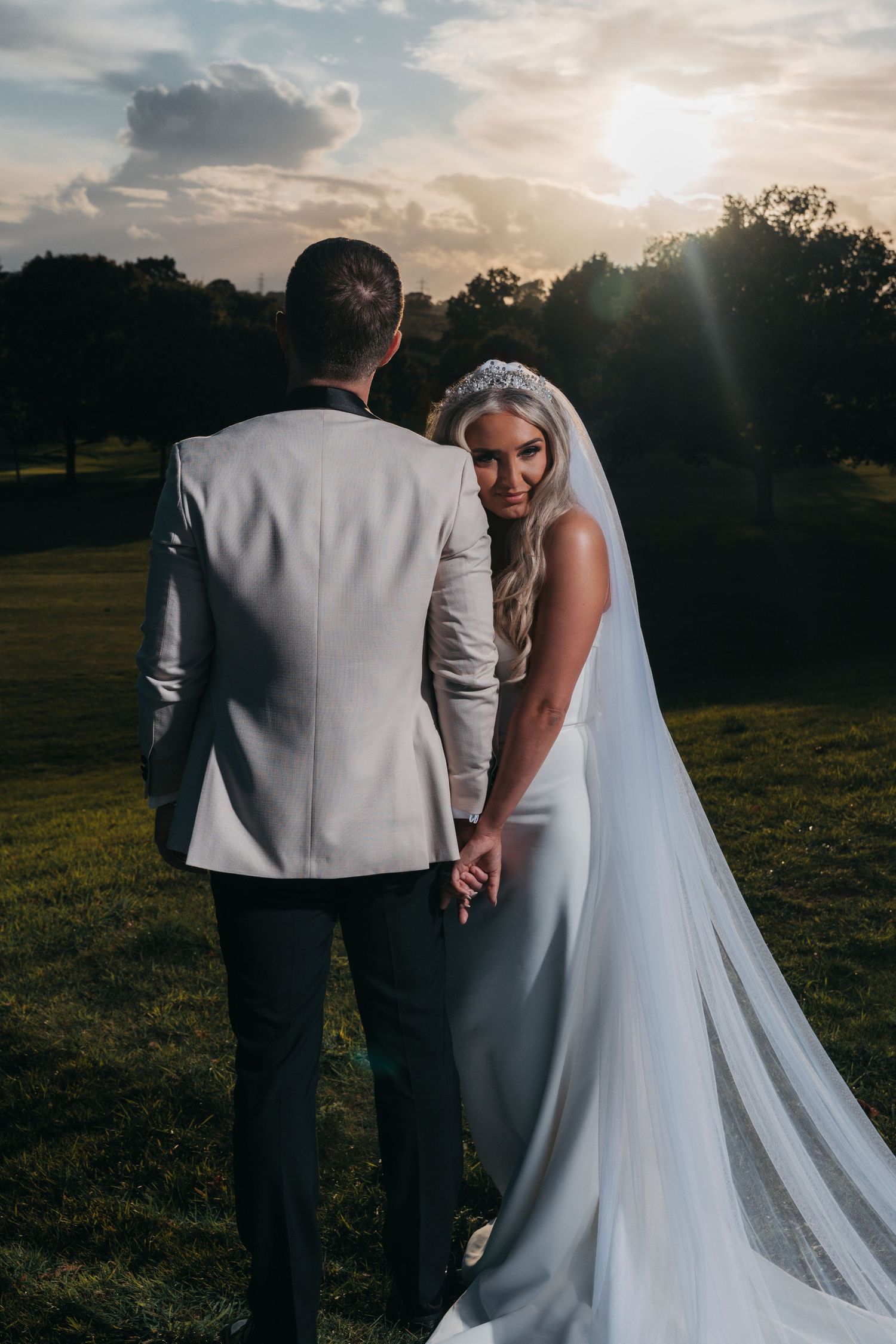 A bride in a long veil and a groom in a light jacket stand together on a grassy hillside at sunset.