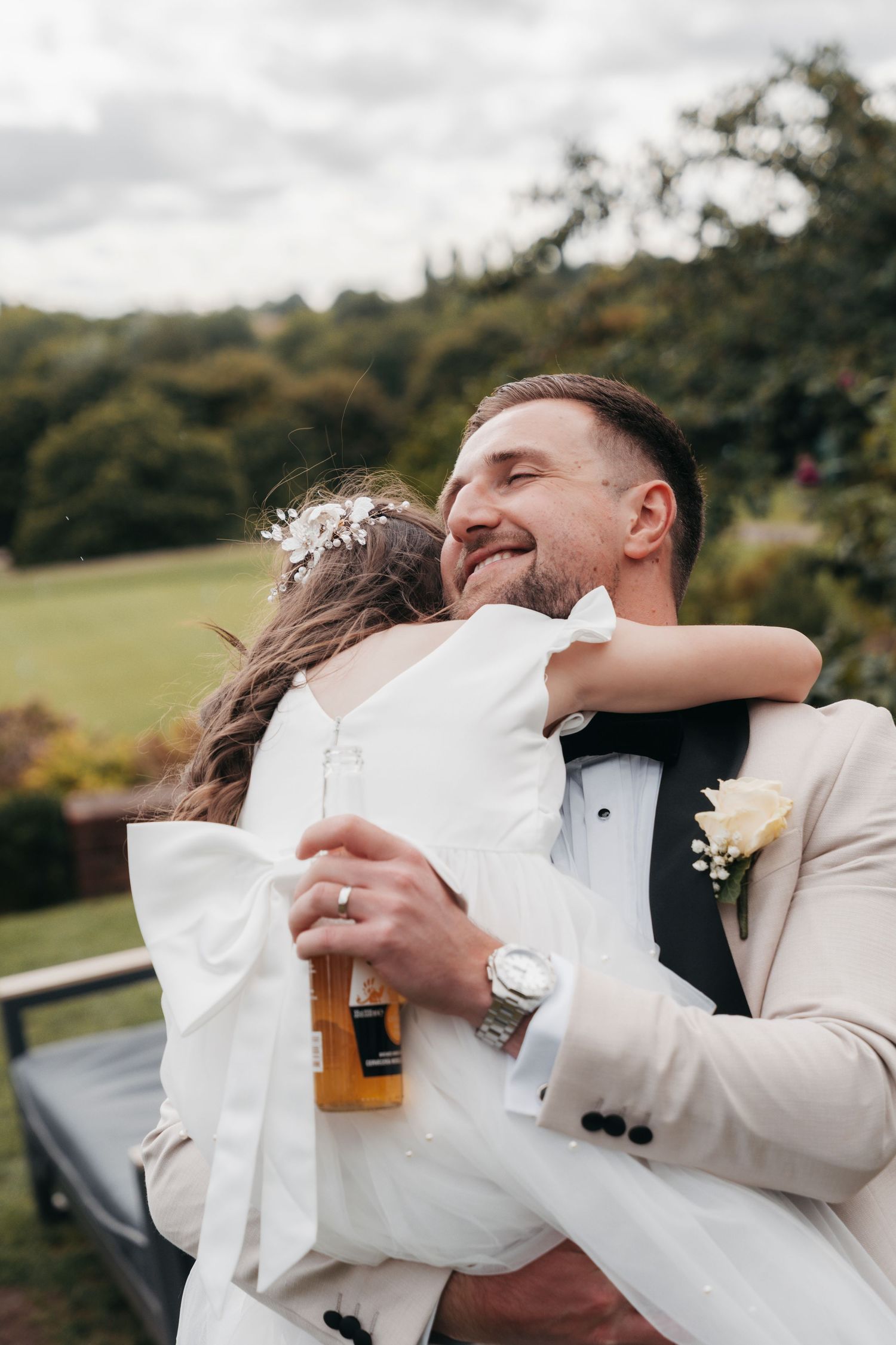 Smiling groom in a beige suit hugs a flower girl in a white dress while holding a drink outdoors at a wedding.