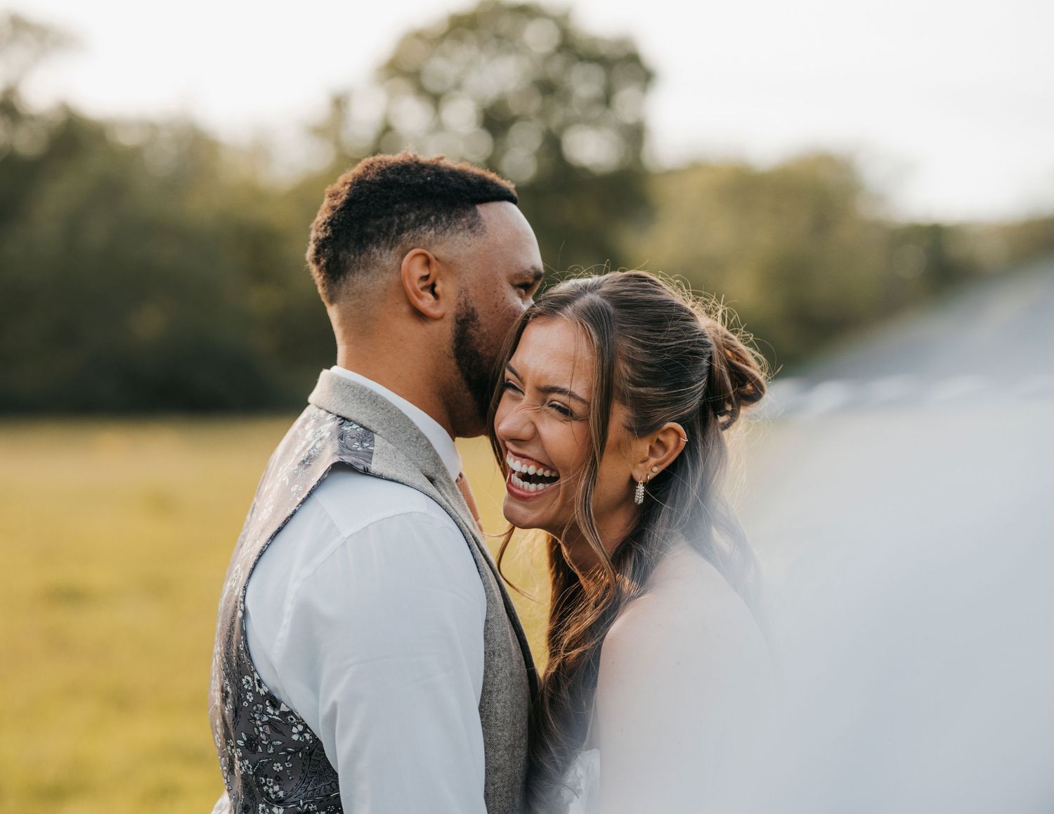 Groom kisses his laughing bride on the forehead as they embrace in a sunlit grassy field at their wedding.