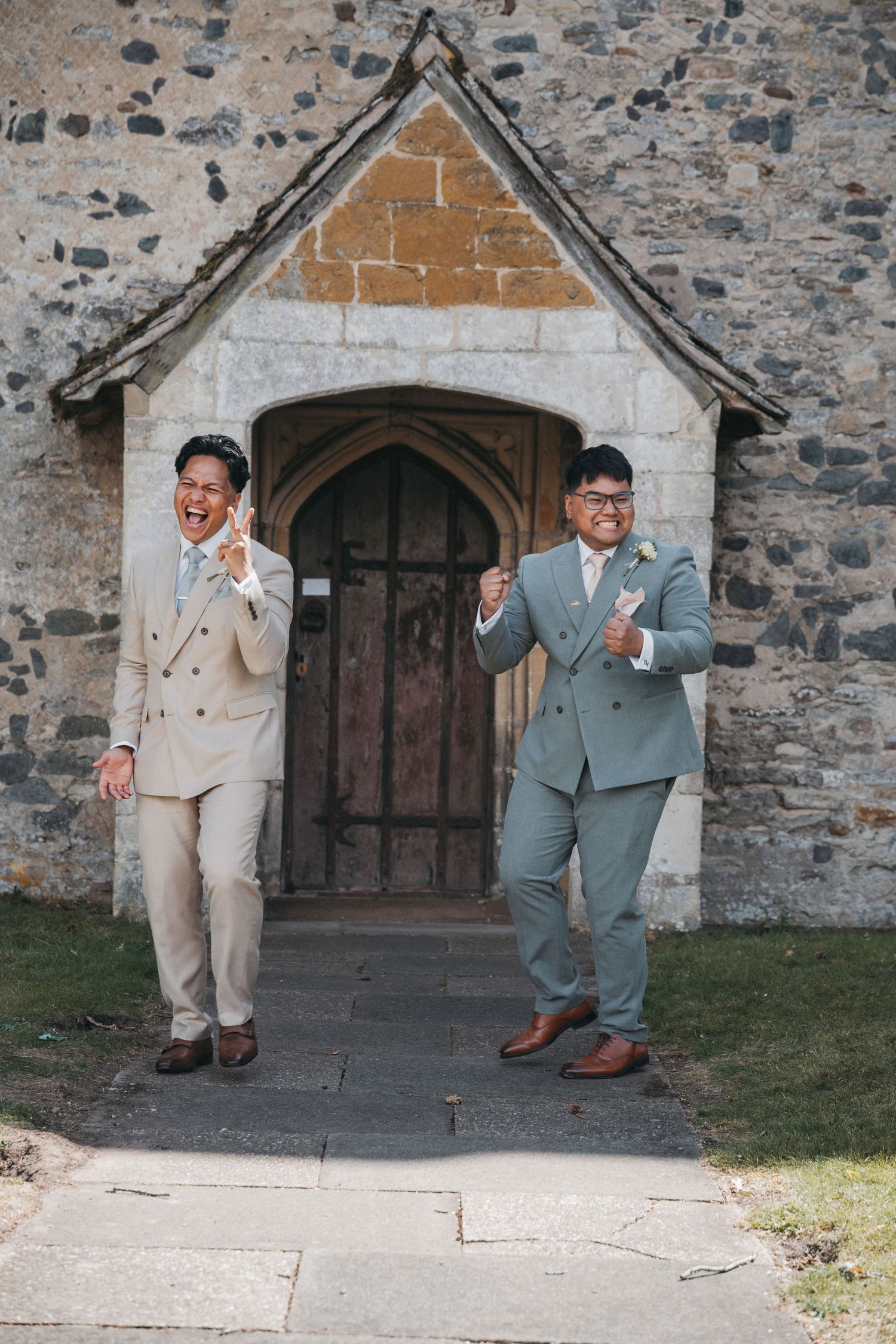 Two grooms in light suits cheer and celebrate with joy as they exit a historic stone church after their wedding.