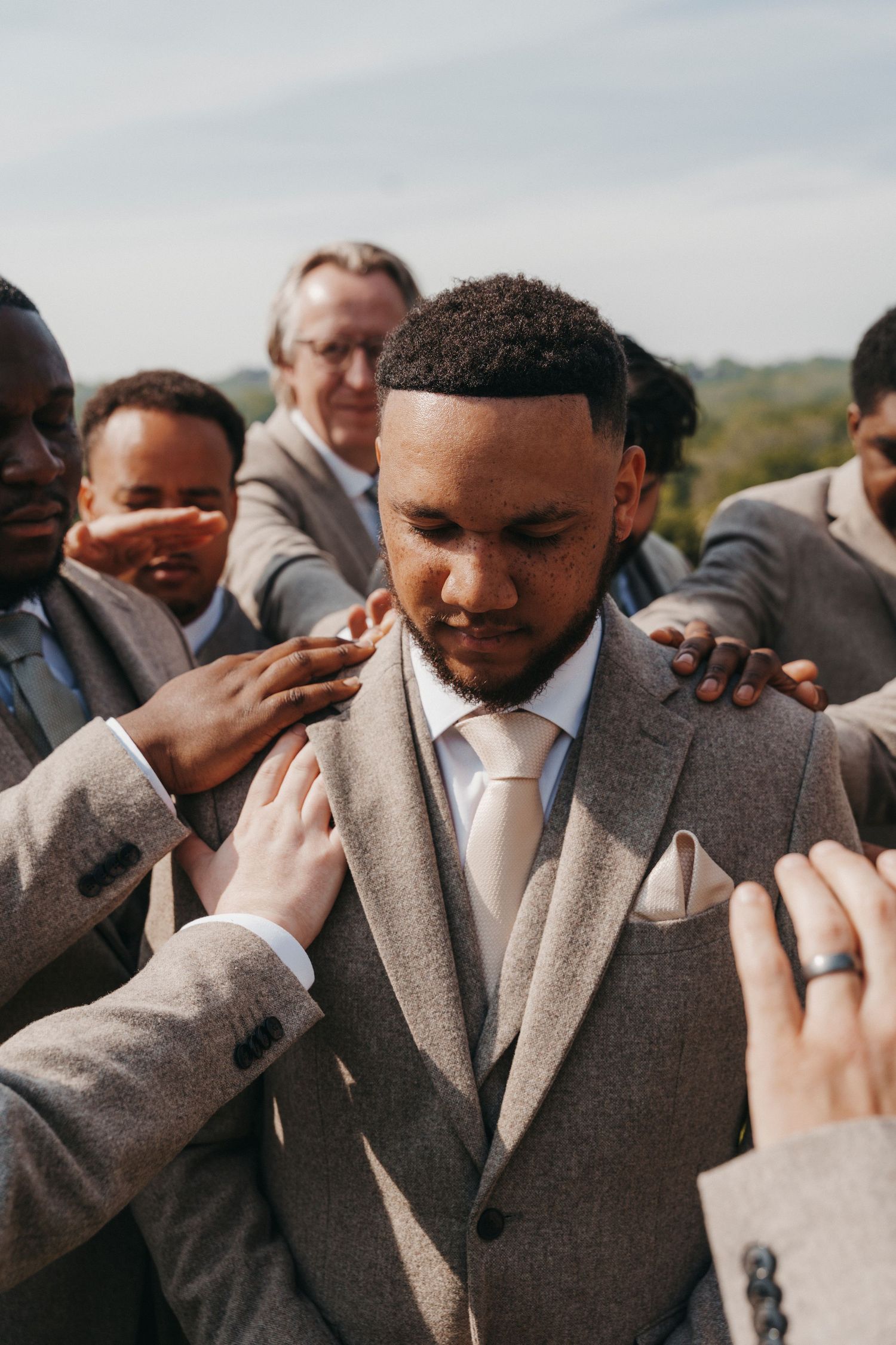Groomsmen and older men place their hands on a groom's shoulders in a heartfelt blessing at an outdoor wedding.