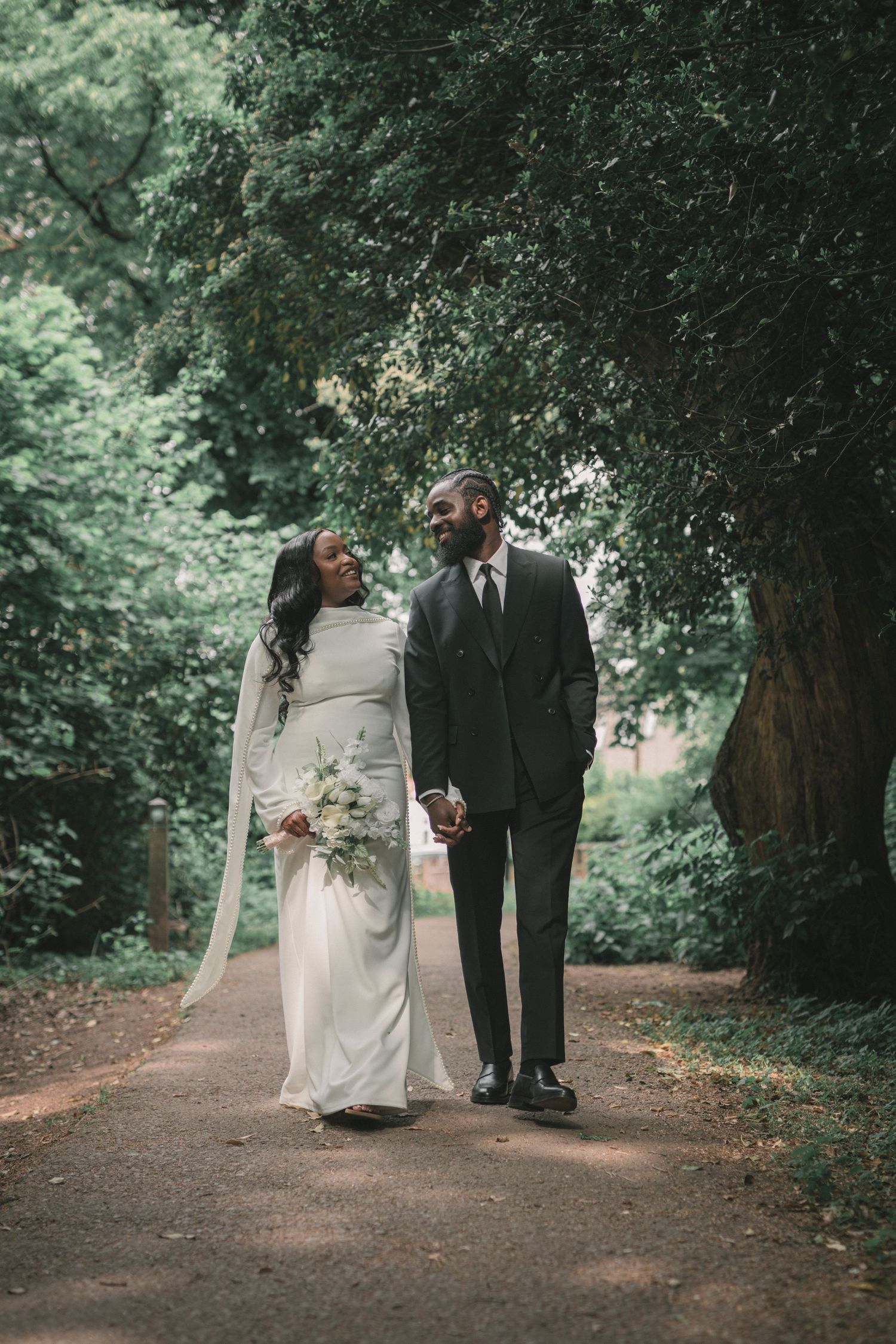 Bride in a white gown holding a bouquet walks hand in hand with her groom along a tree-lined path, smiling at each other.