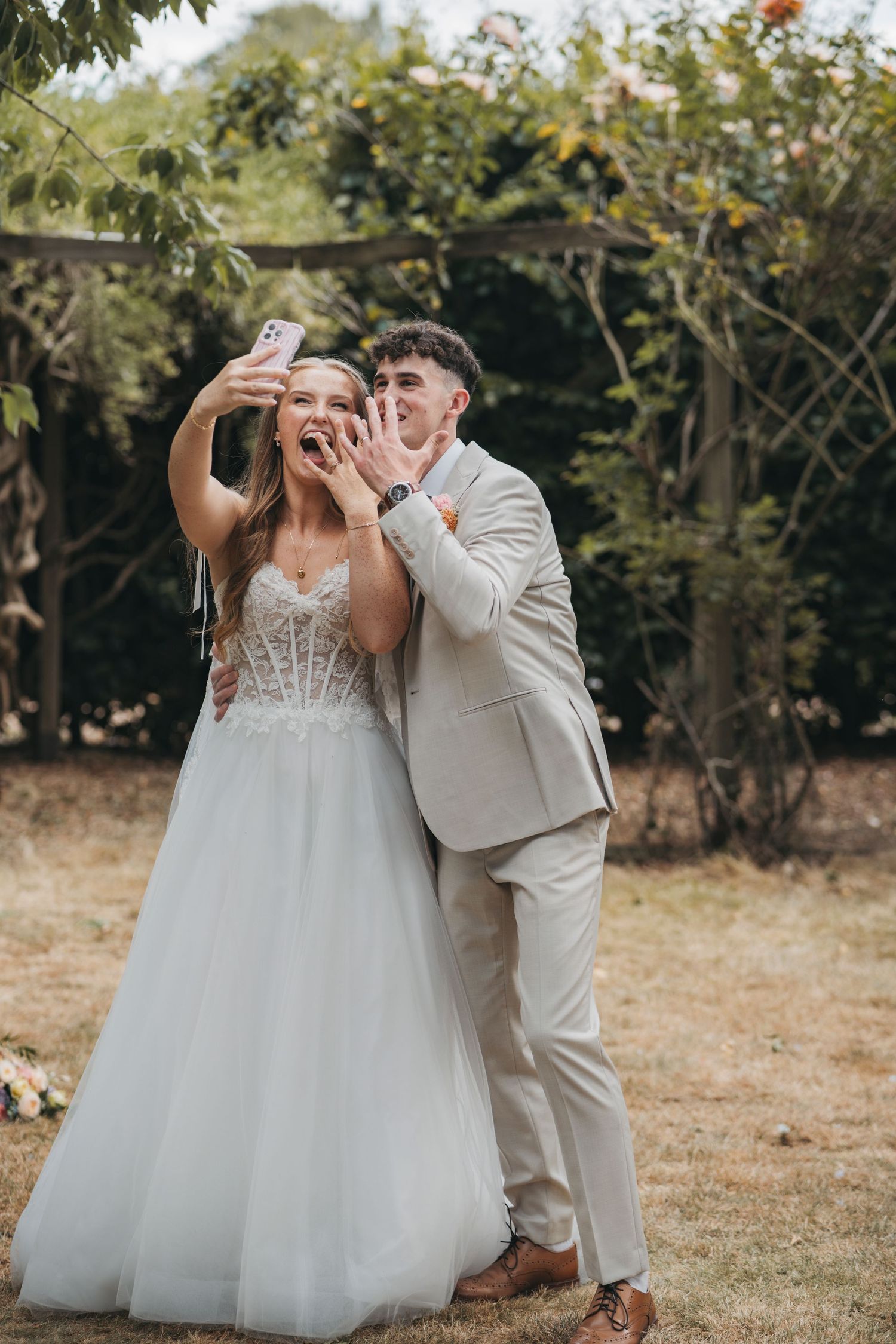 Joyful bride in a tulle gown and groom in a beige suit show their rings and take a selfie in a leafy garden.