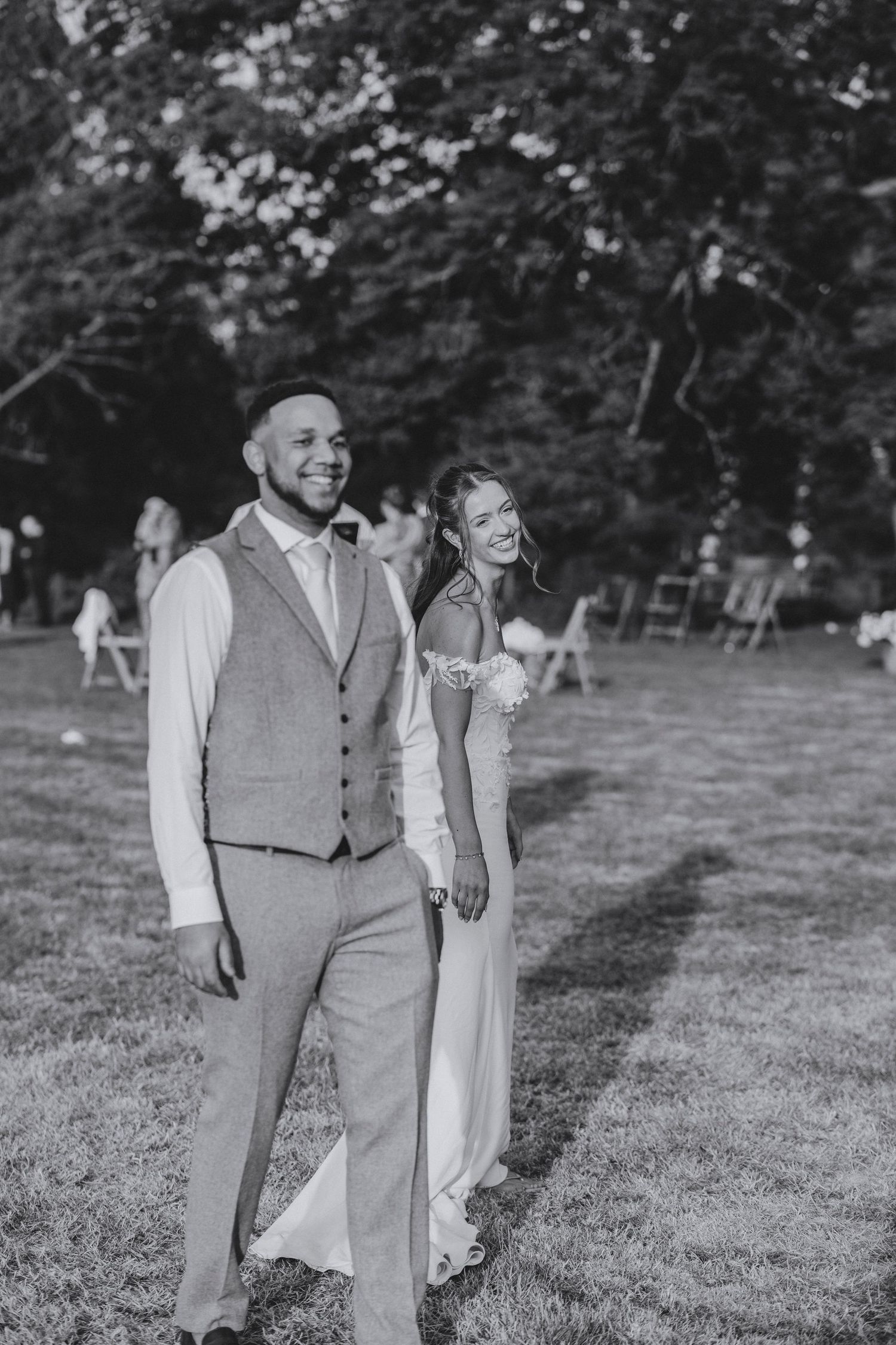 Black and white photo of a smiling groom in a waistcoat walking hand in hand with his bride across a lawn.