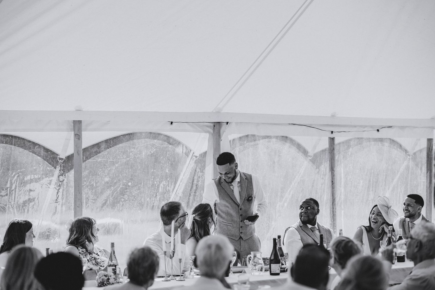 Black and white photo of a groom standing to give a speech to seated guests at a long table inside a wedding marquee.