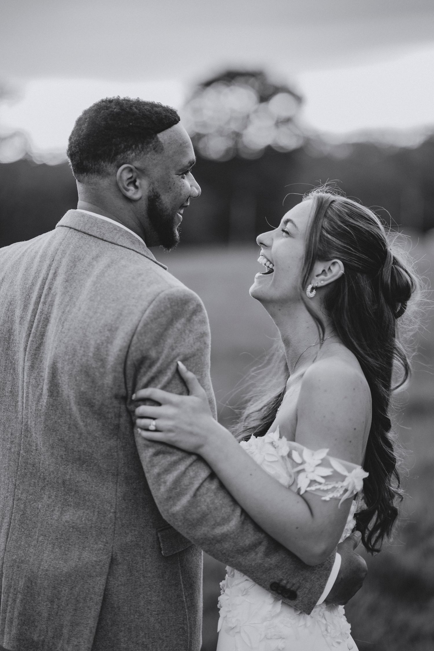 A bride and groom share a joyful laugh while embracing in a black and white outdoor wedding portrait.