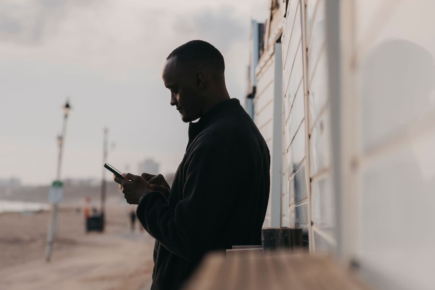 Silhouetted man in a dark jacket looks down at his smartphone while standing beside a wooden beach hut at dusk.