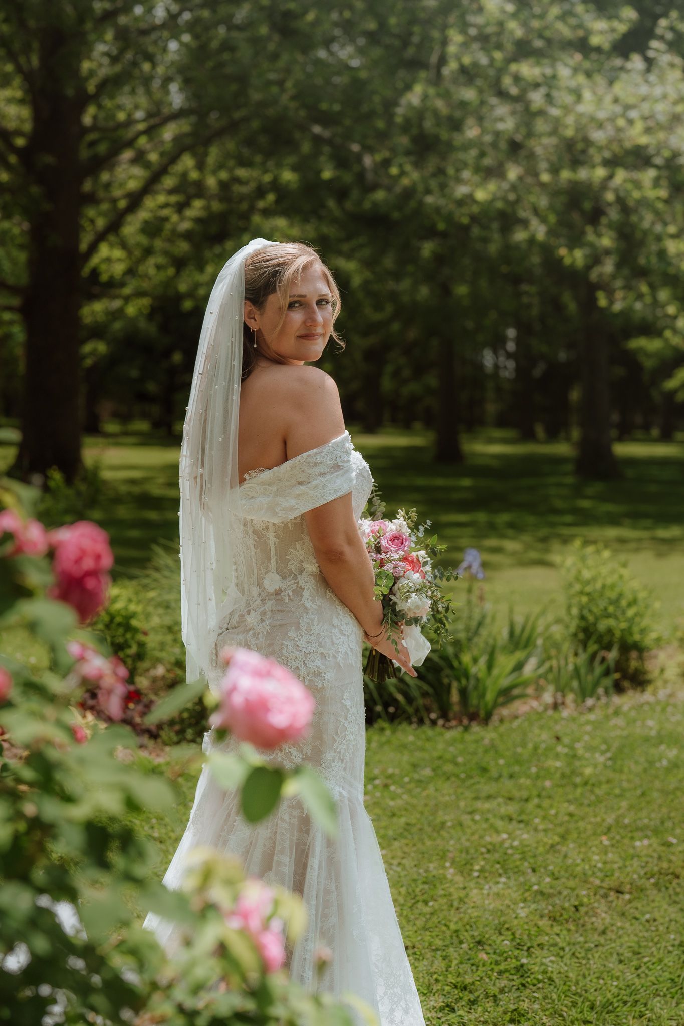 Bride in lace off-shoulder gown and veil holds bouquet among pink roses in lush green garden.
