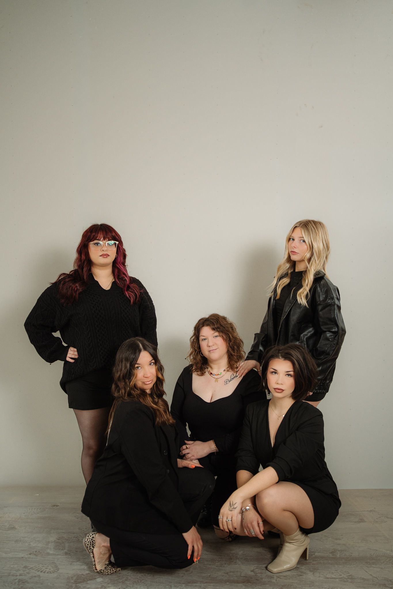 Five women in black outfits pose together against a light gray wall in a stylish group portrait.
