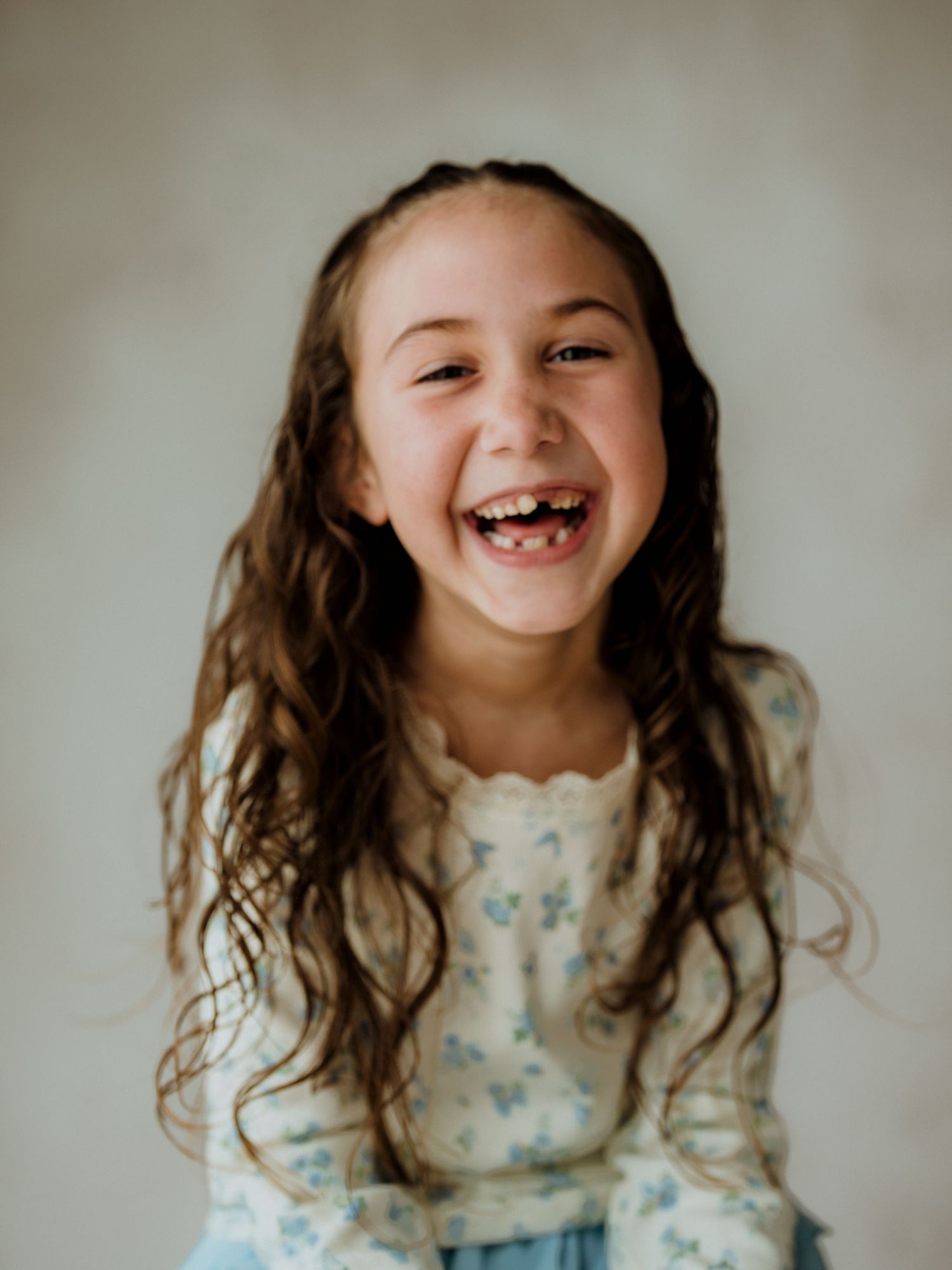 A joyful young girl with long curly brown hair laughs brightly, wearing a floral top against a neutral background.