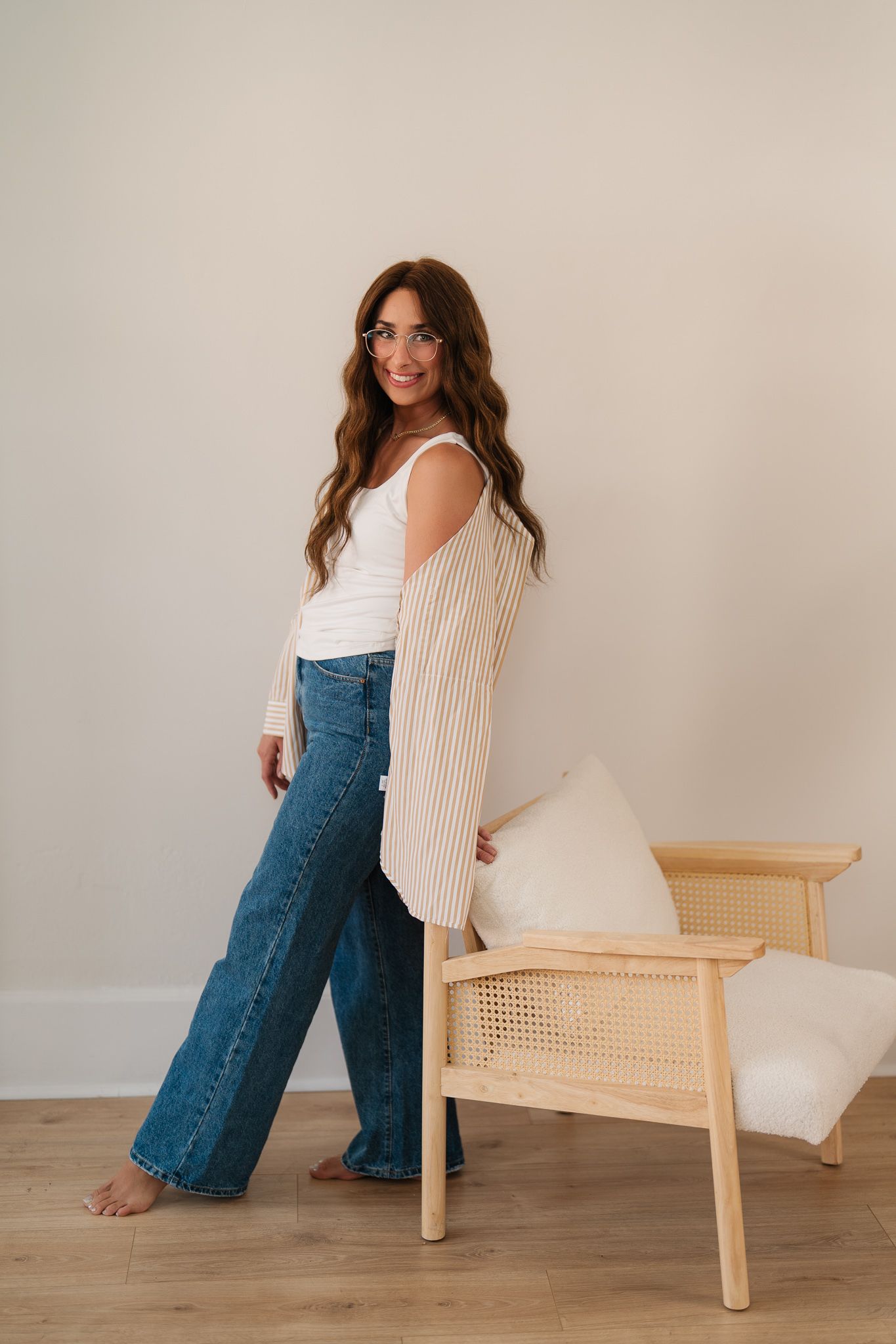 Woman in wide-leg jeans, white tank, and striped cardigan posing beside a rattan chair in a minimalist room.