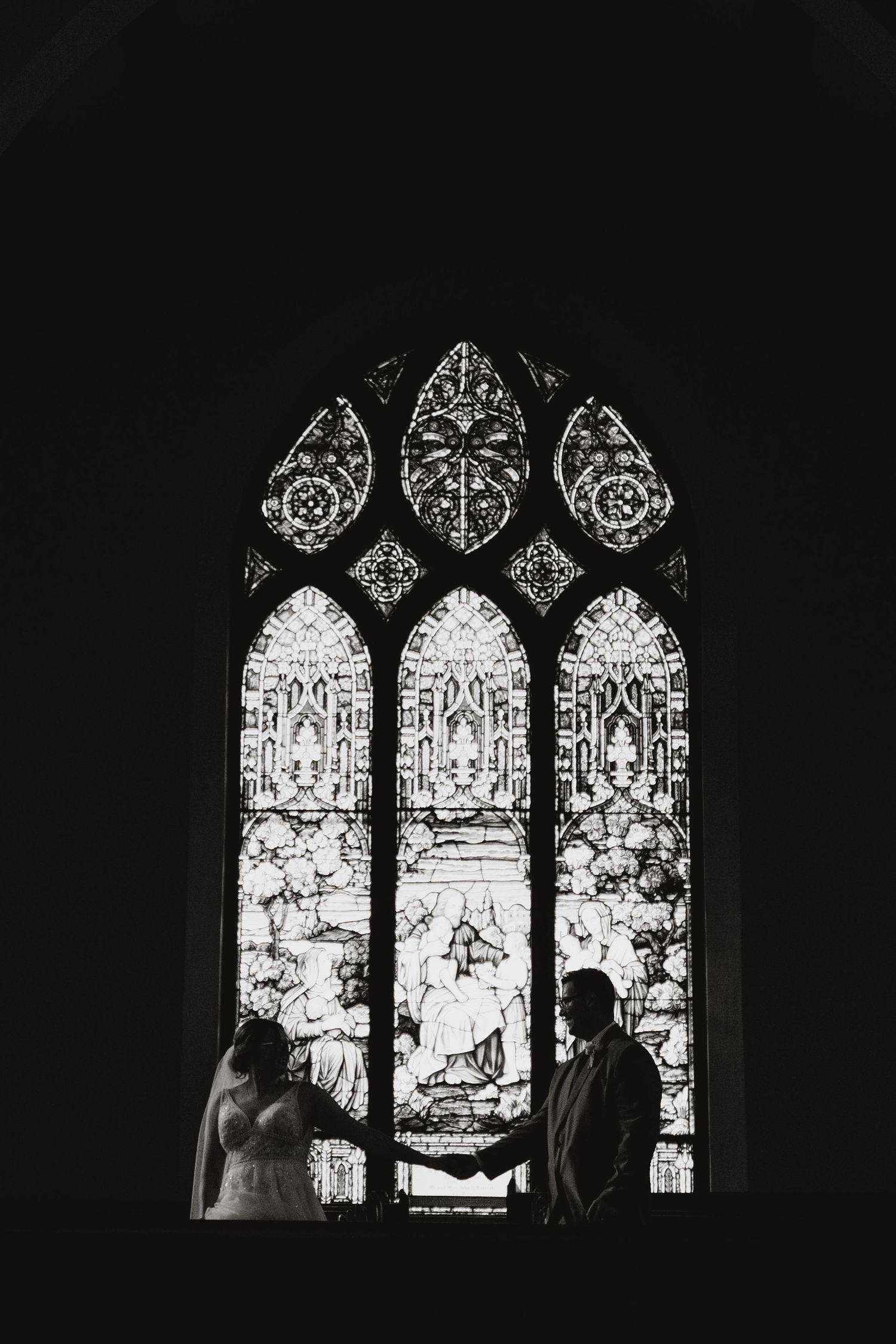 A silhouetted couple stands before an ornate stained glass church window. Tiffin, Ohio Wedding Venue