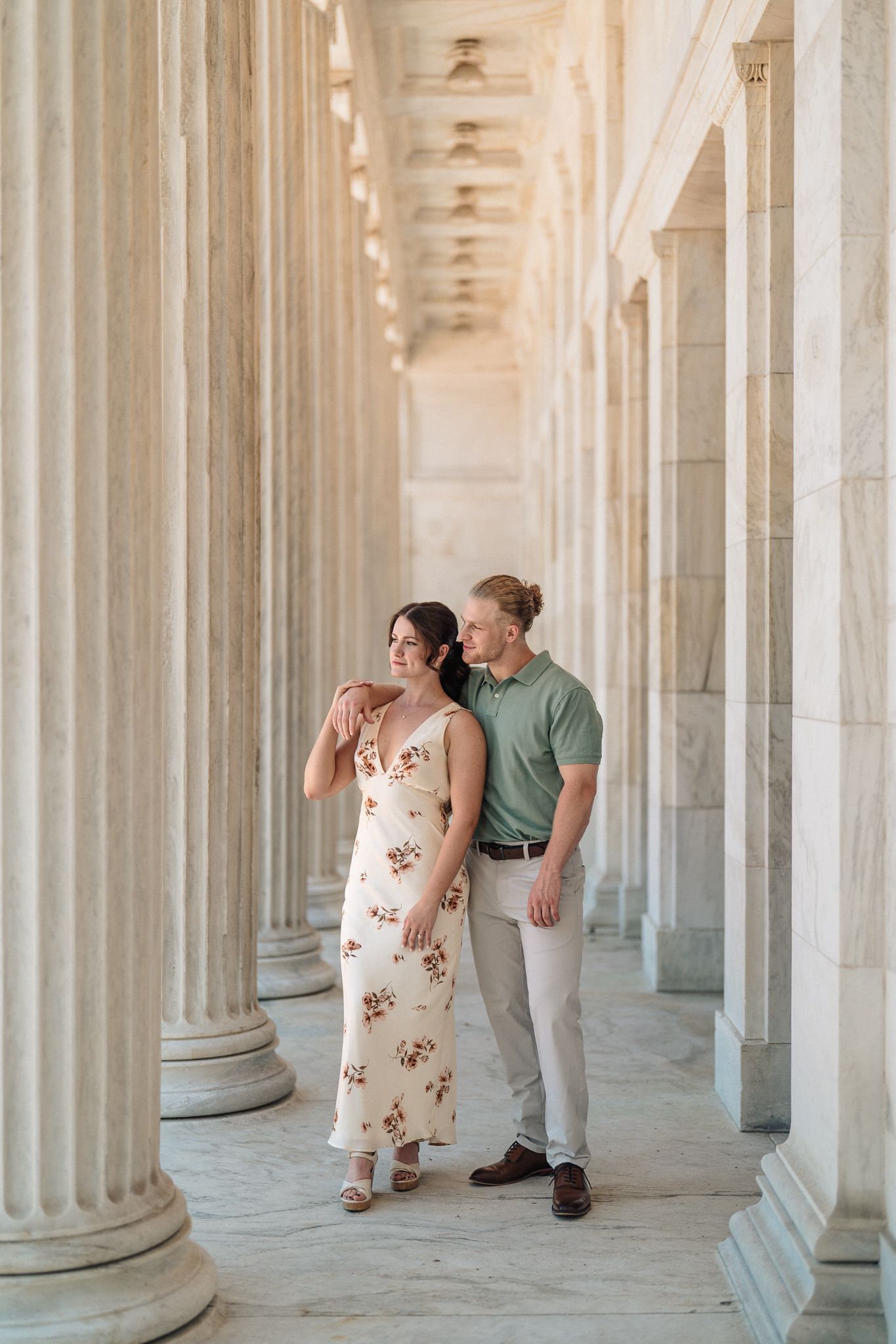 Couple posing among tall classical columns, woman in floral dress and man in green polo shirt, elegant architectural setting.