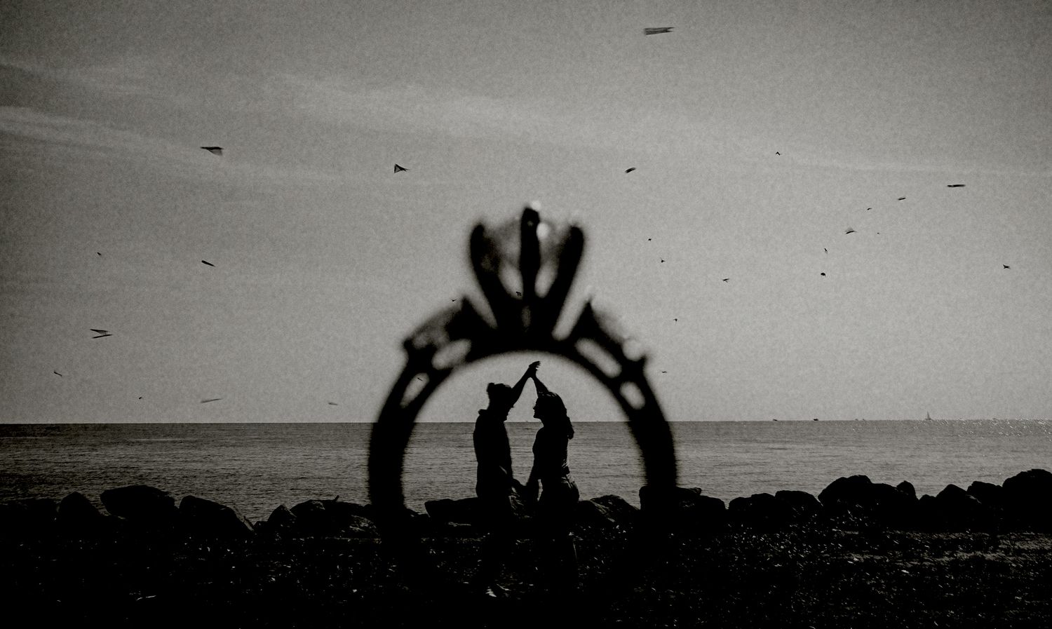 Silhouette of couple framed by engagement ring shadow on rocky beach at dusk.