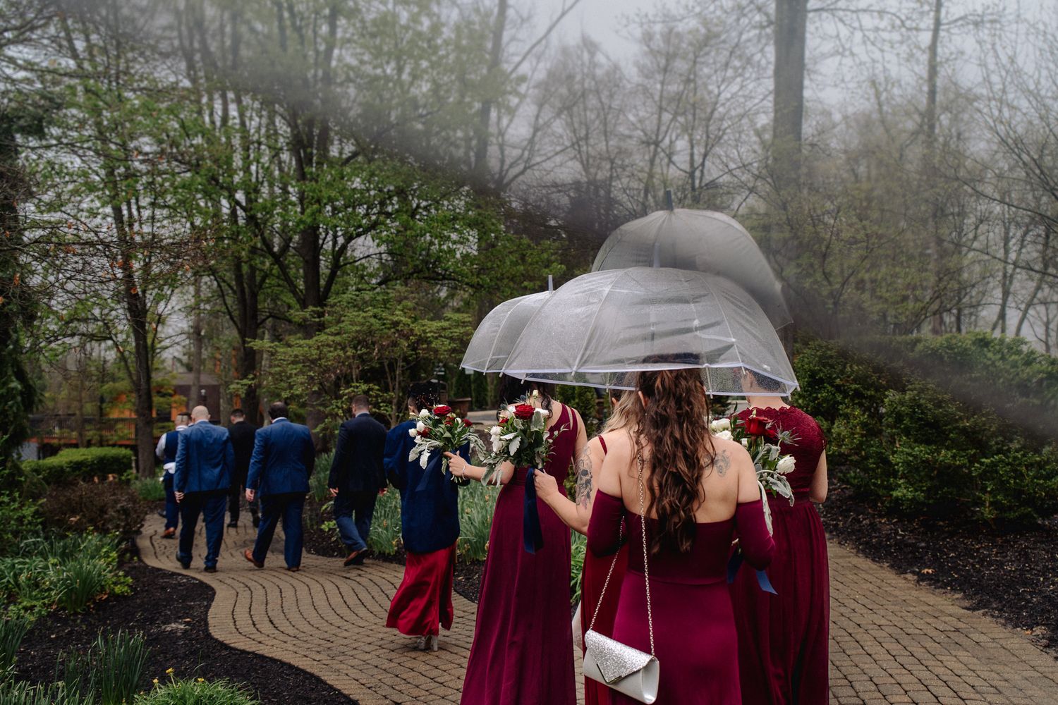 Bridesmaids in burgundy dresses holding clear umbrellas and red bouquets walk toward groomsmen on a rainy garden wedding day.
