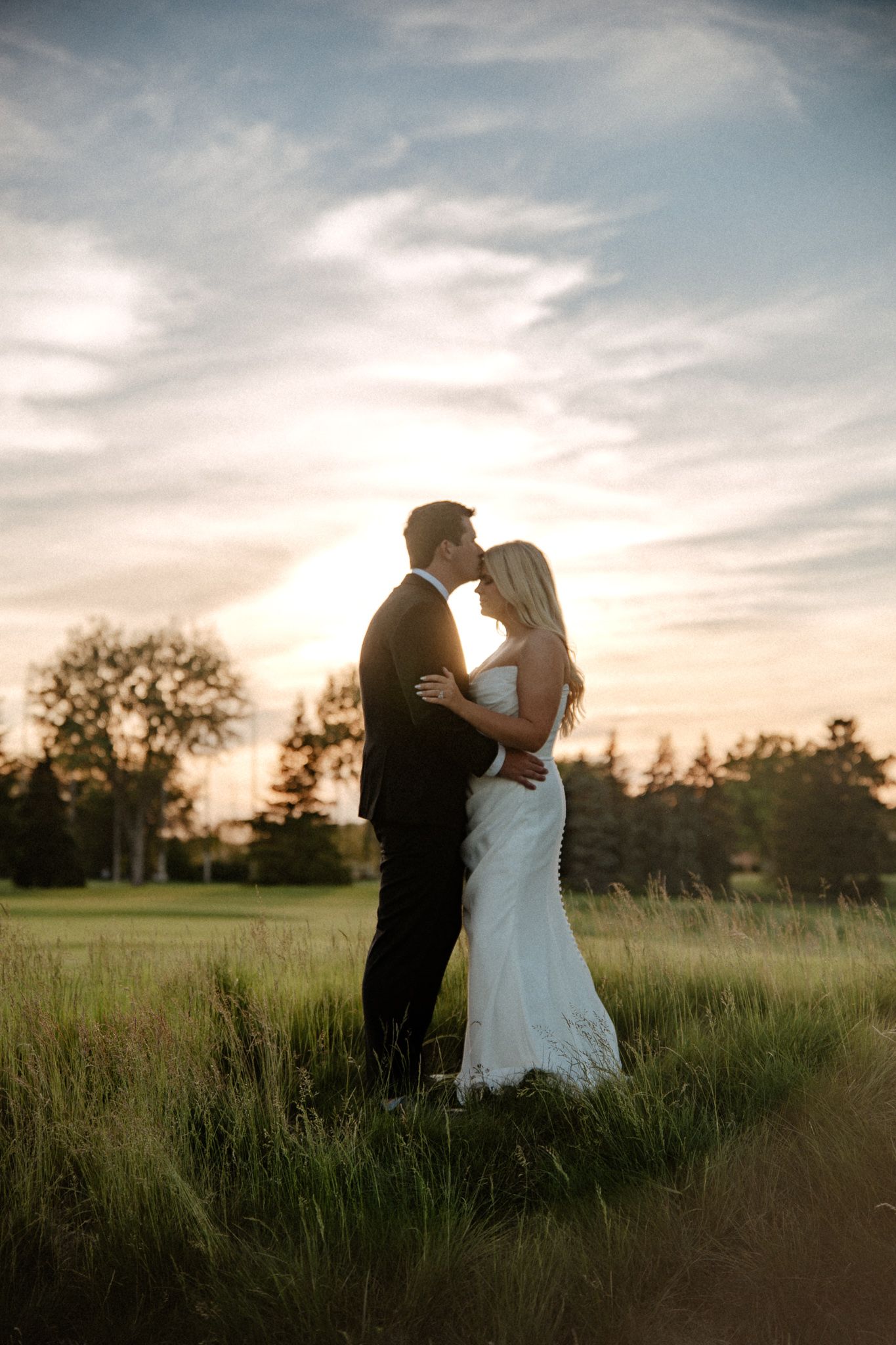 Bride and groom share a romantic kiss at sunset in a lush green field, with trees and a dramatic sky behind them.