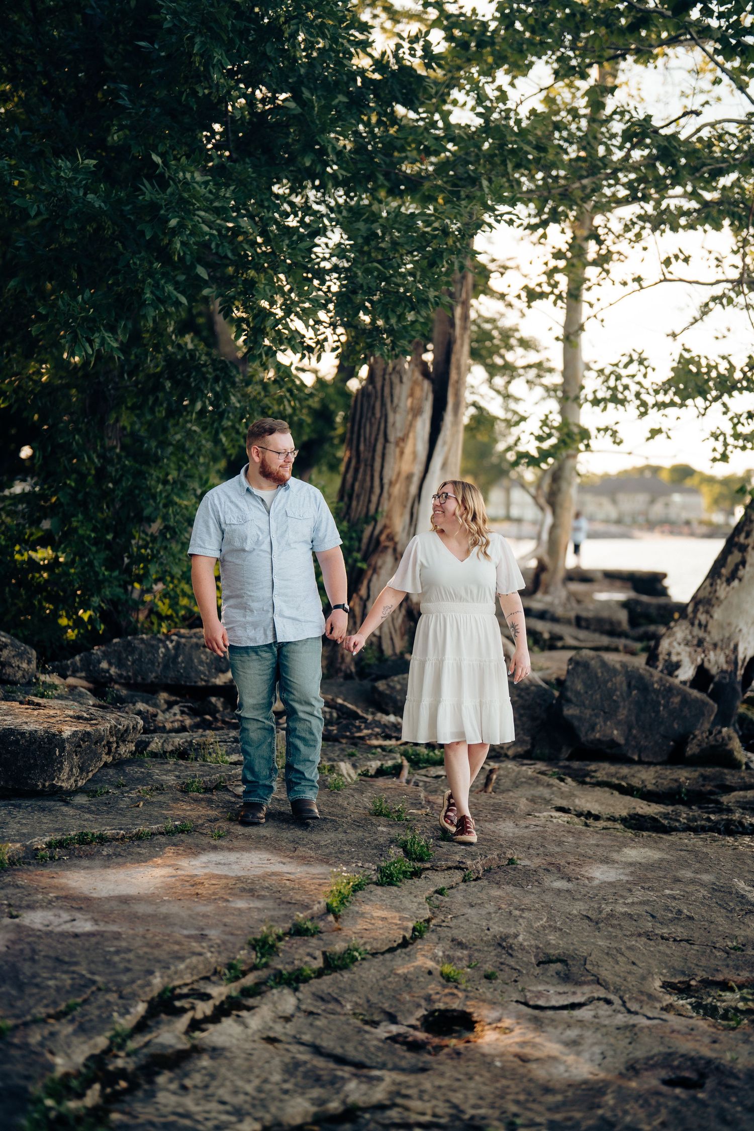 A couple holds hands walking on rocky terrain near a waterfront, surrounded by lush trees in warm golden light.