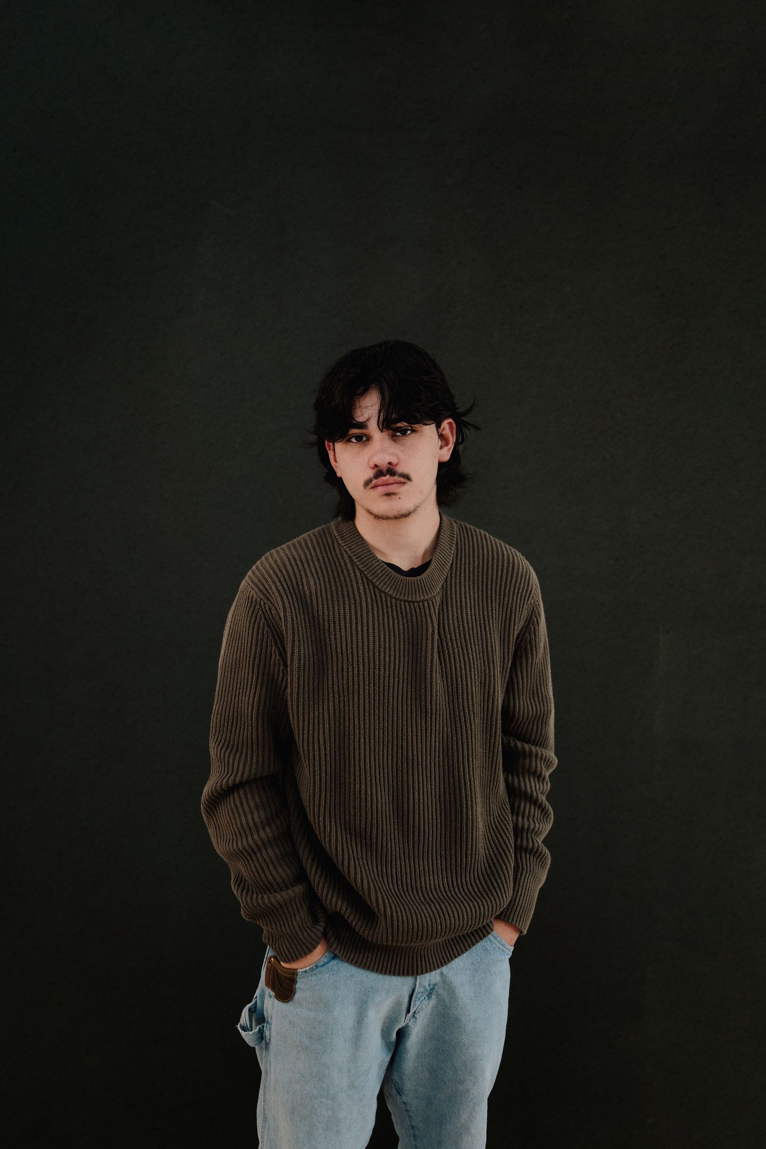 Young man with mustache wearing olive ribbed sweater and light jeans, standing against dark background.