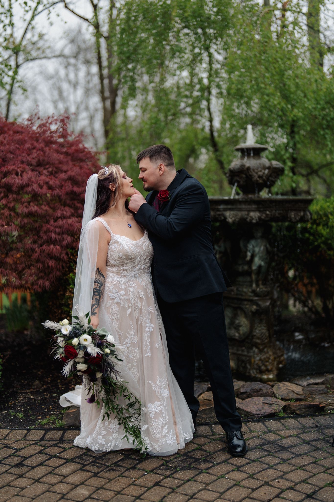 A bride and groom share a romantic moment in a garden, she holds a bouquet in a lace gown, he wears a black suit.