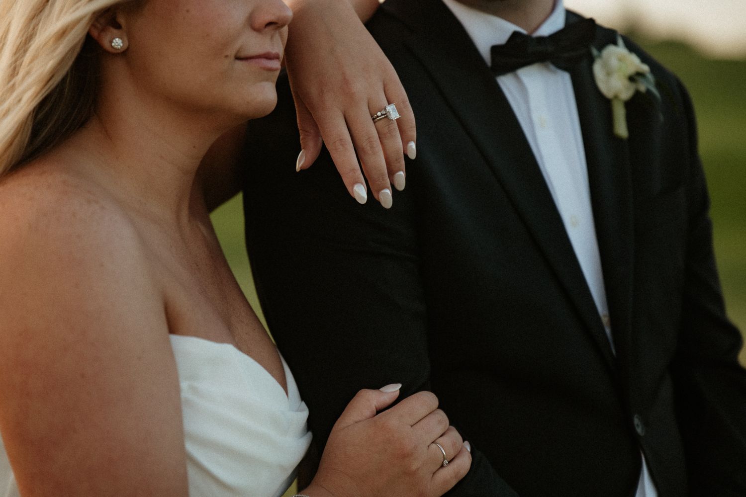 Bride in strapless white gown showing diamond engagement ring while holding groom's arm in black tuxedo outdoors.