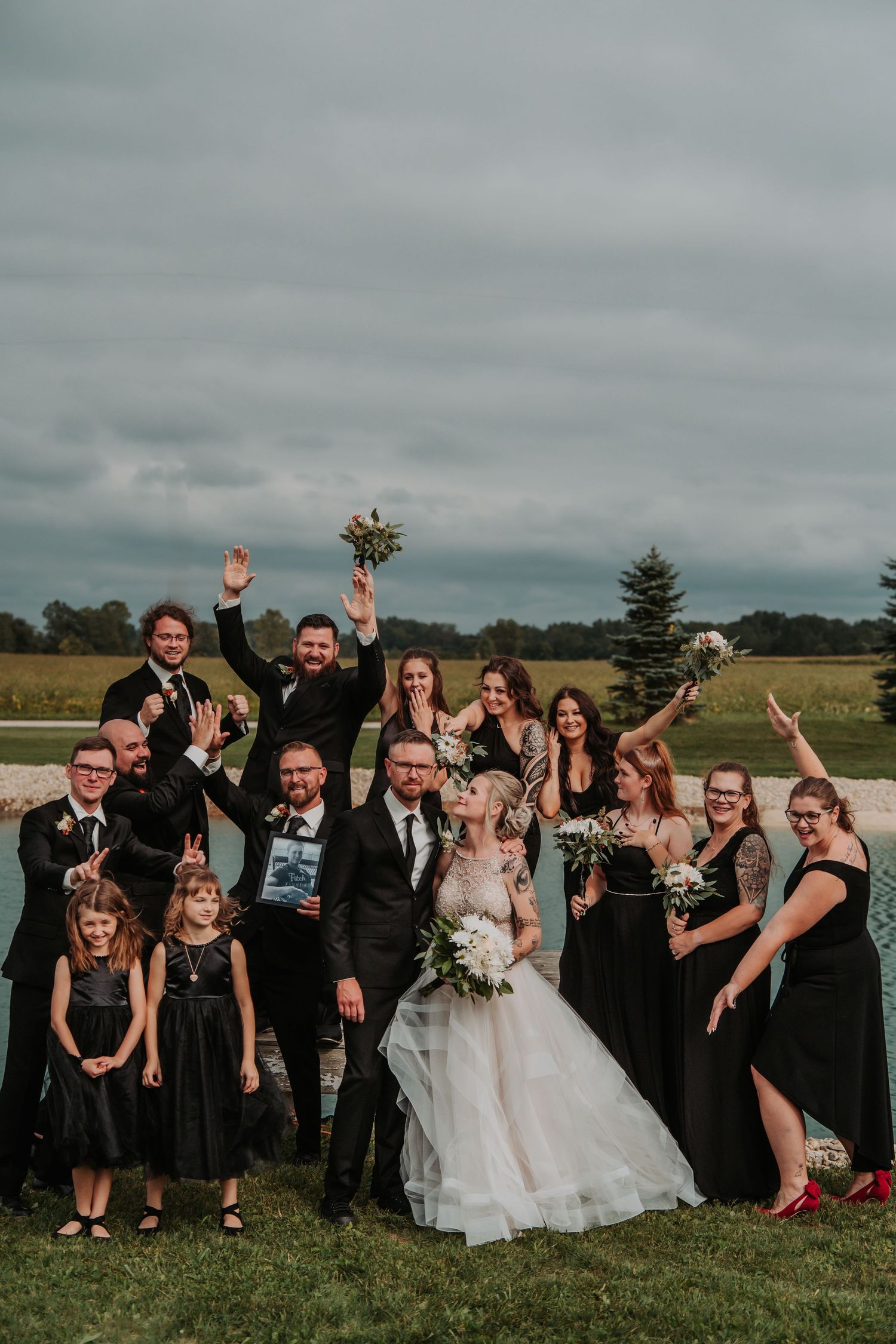 Energetic wedding party in black attire posing together on a grassy field. Tiffin, Ohio Wedding Venue