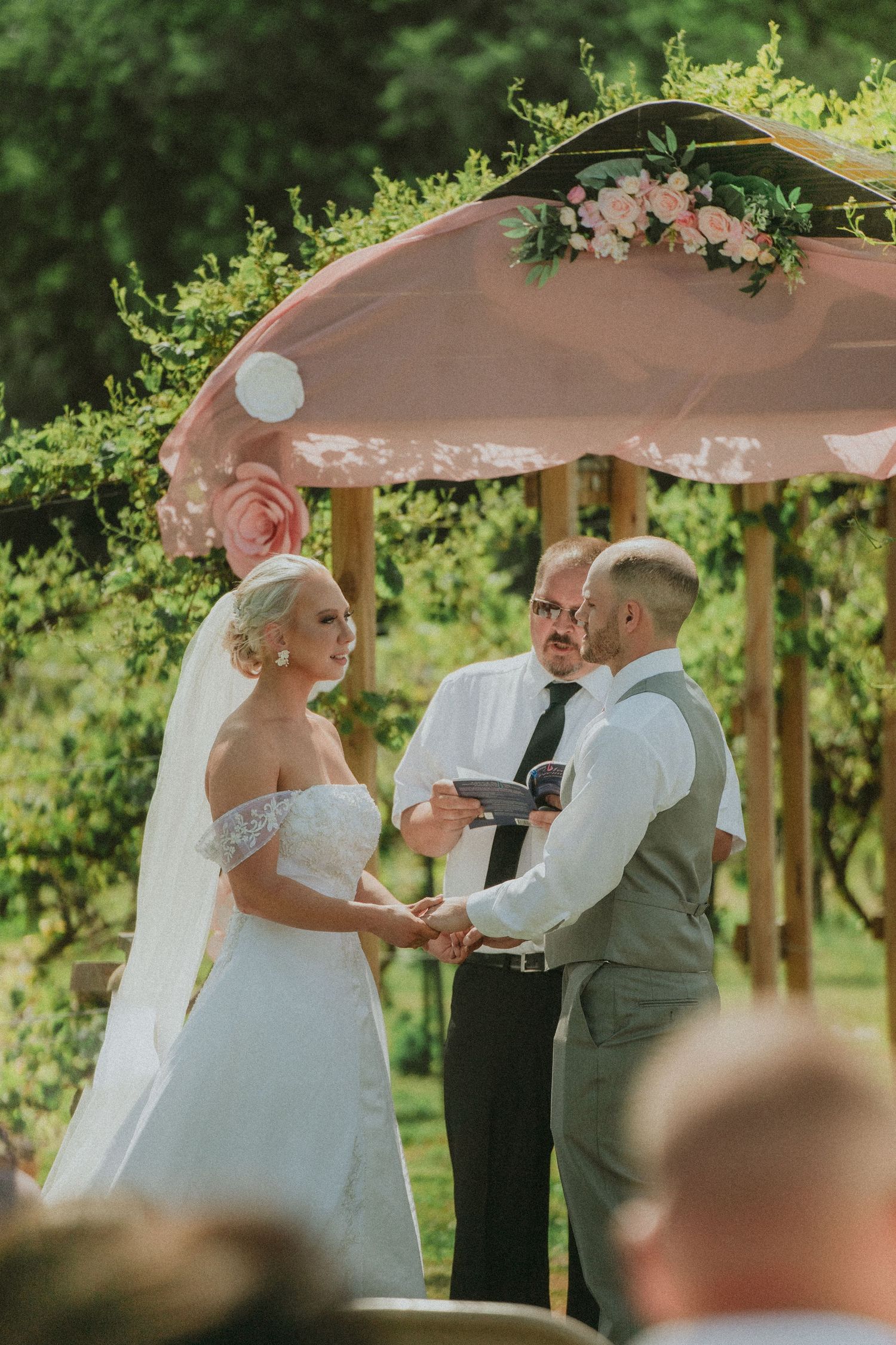 An outdoor wedding ceremony under a pink-draped arch decorated with fresh flowers.  Clyde, Ohio copper whale winery