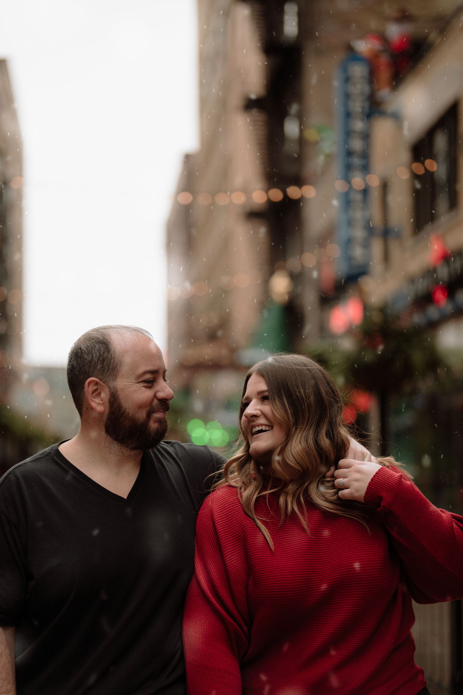 A couple laughs together during a winter urban photoshoot with city lights in the background. Downtown Cleveland, Ohio