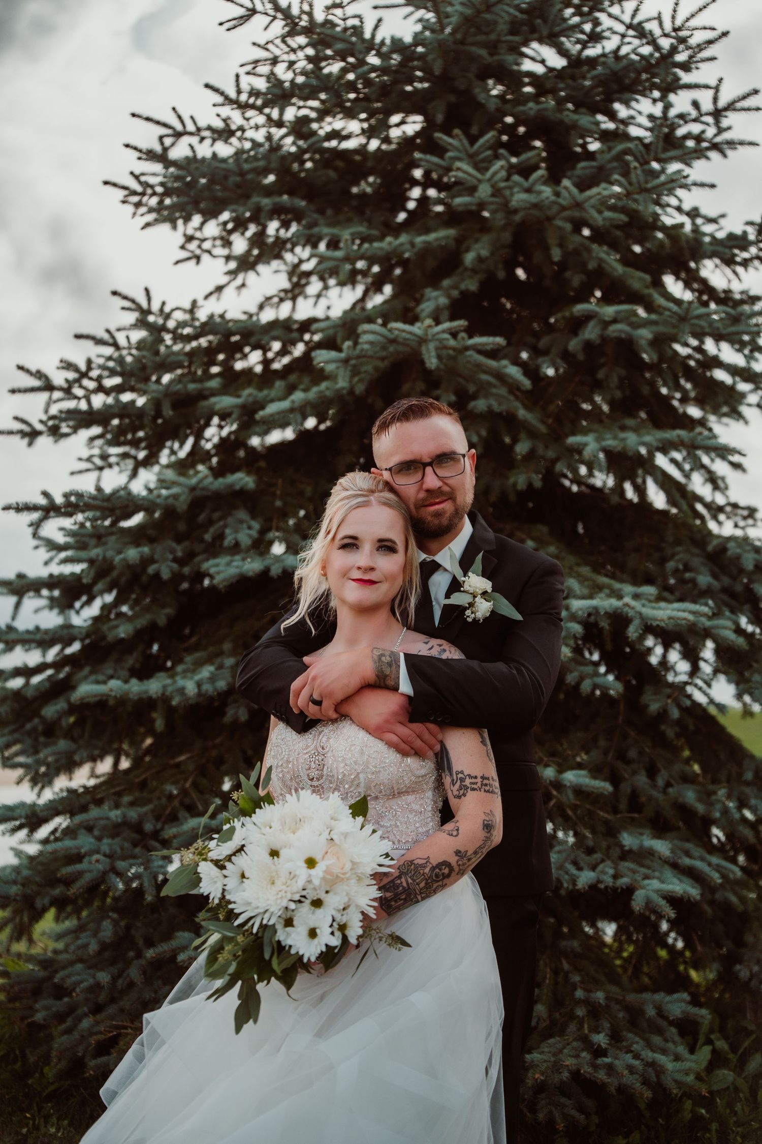 Wedding couple poses in front of evergreen trees with white floral bouquet. Tiffin, Ohio Wedding Venue