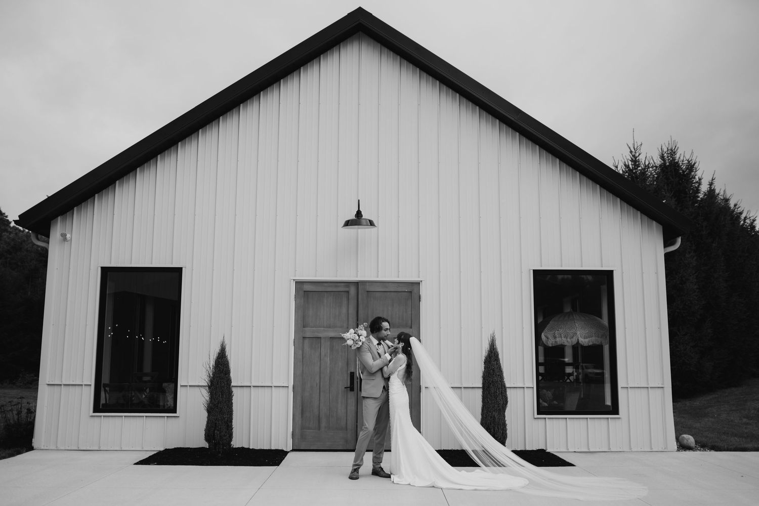 A black and white photo of a couple standing in front of a modern white barn with cypress trees. Wooster Ohio The Wellspring Wedding Venue