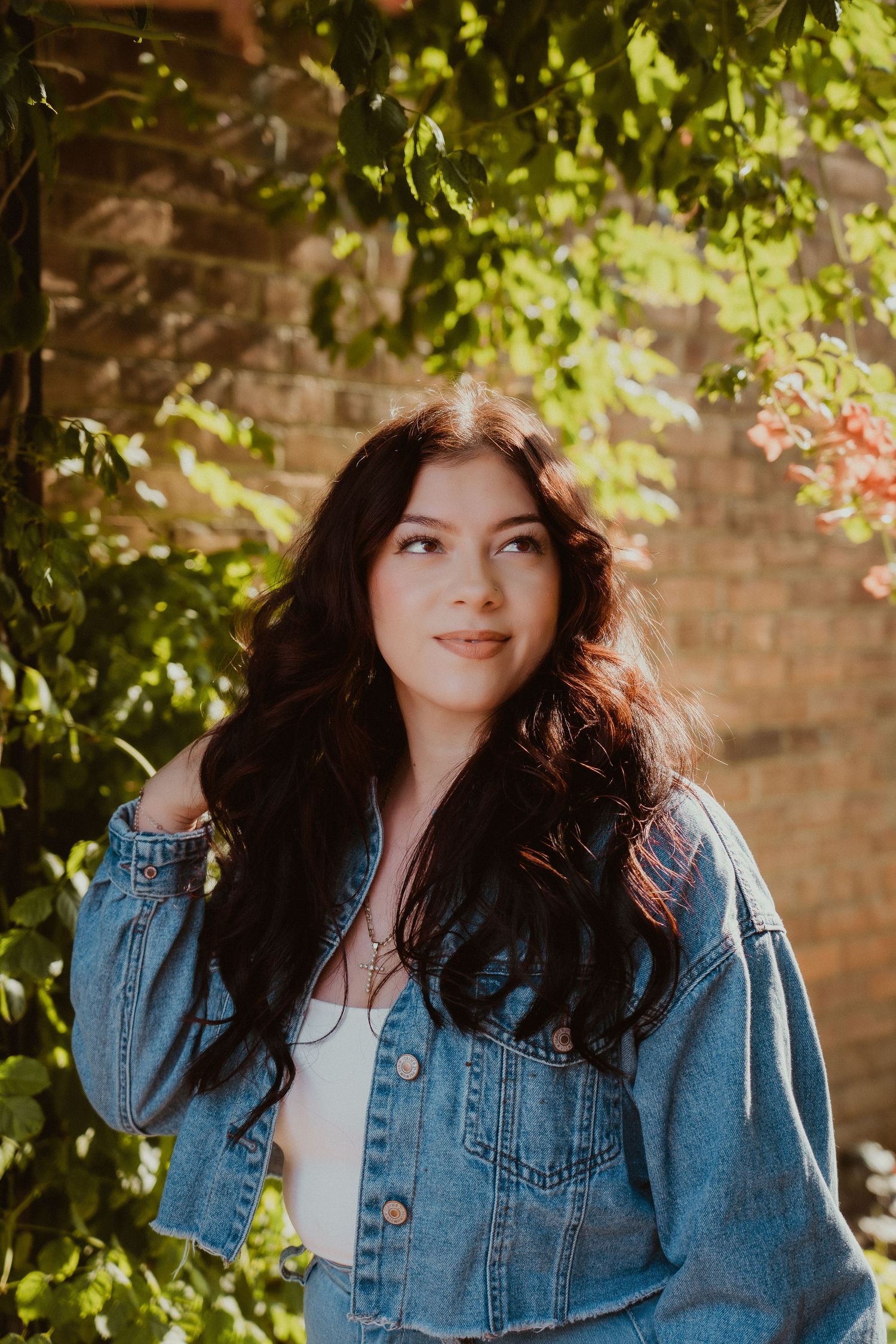 Woman in denim jacket poses against leafy backdrop in natural outdoor lighting. Fremont, Ohio Hair Salon