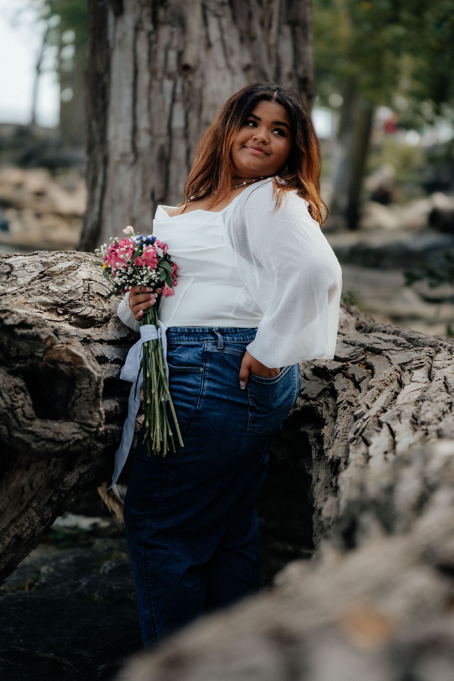A person wearing a white blouse and jeans sits on a fallen tree trunk while holding a bouquet of pink flowers. Marblehead Lighthouse Ohio