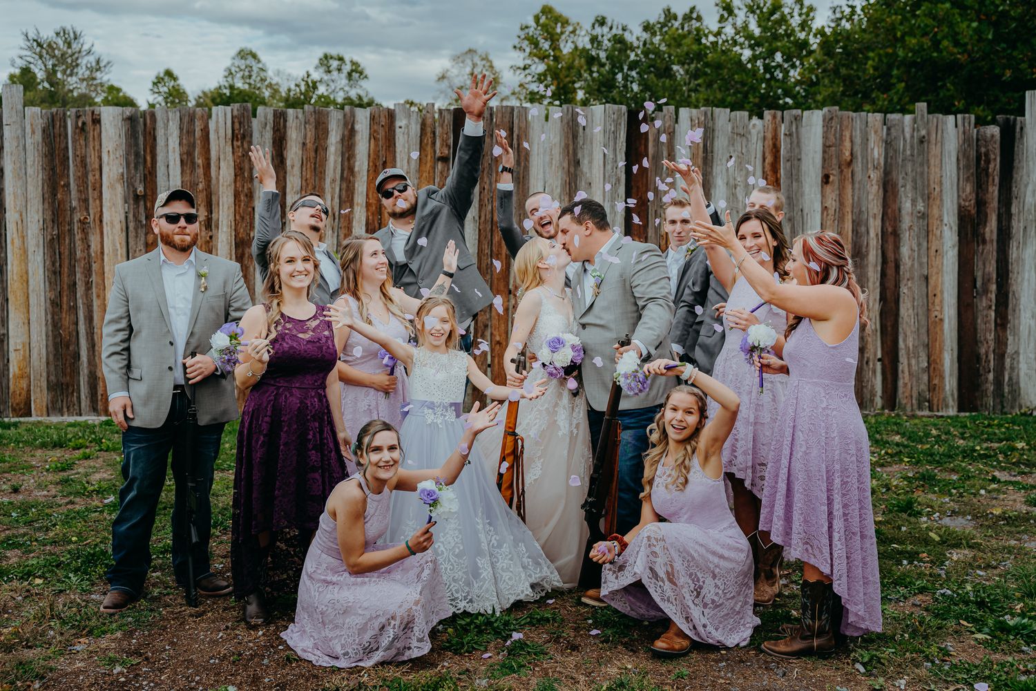 A wedding party poses together in front of a rustic wooden fence wearing purple bridesmaid dresses. Gibsonburg OHIO