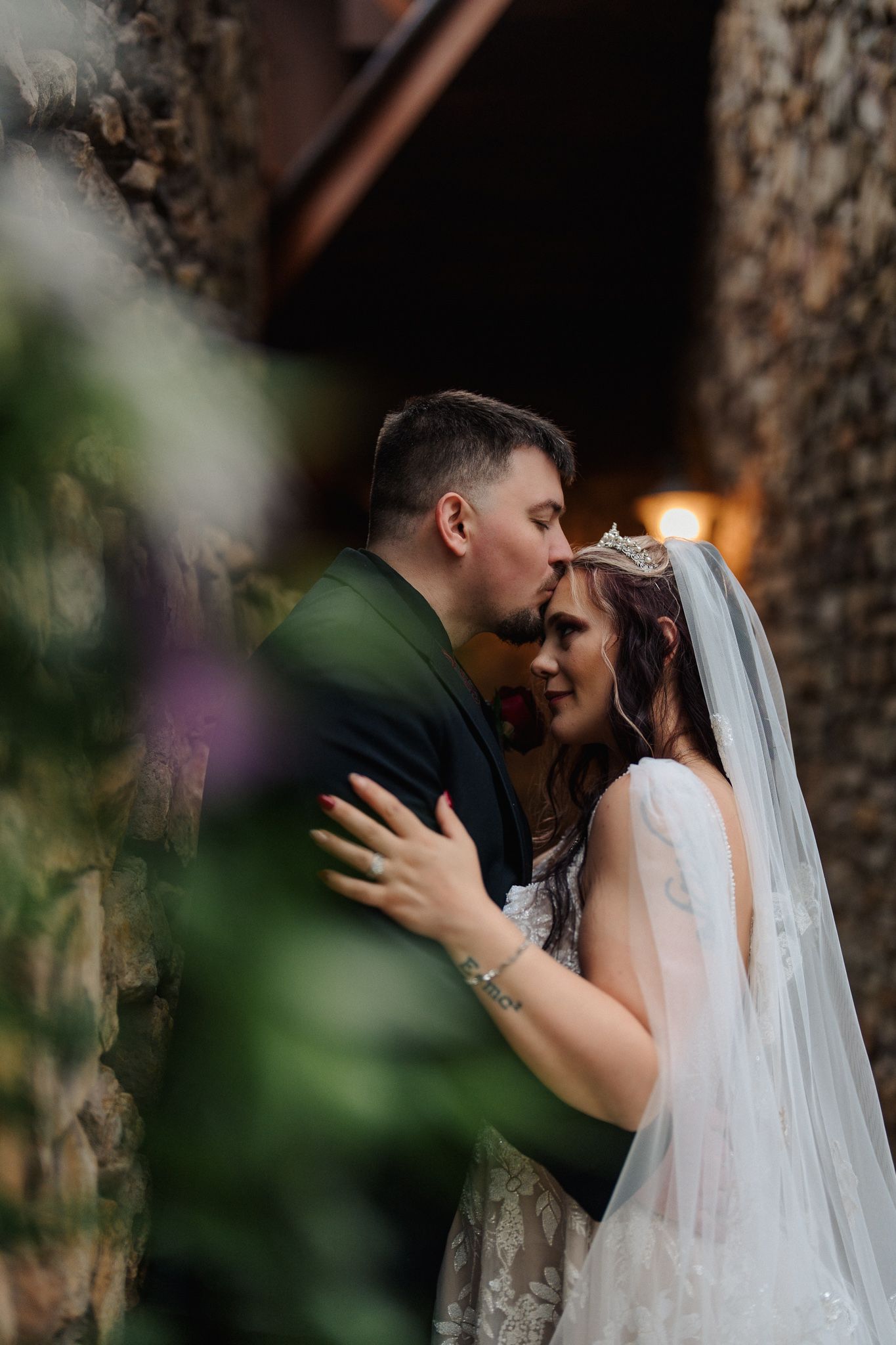 A bride and groom share an intimate moment, embracing near a stone wall, she wearing a veil and tiara, he in a dark suit.
