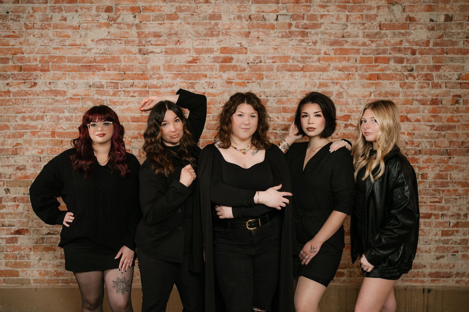 Five women in black outfits pose together against a rustic brick wall for a stylish group portrait.