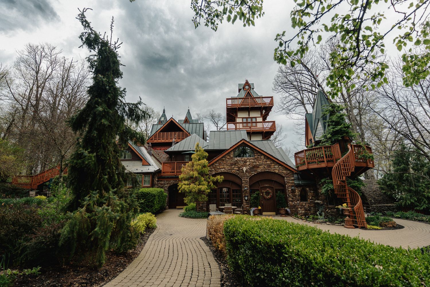 Rustic wooden mansion with stone accents, lush gardens, and brick pathway surrounded by bare trees under cloudy sky.