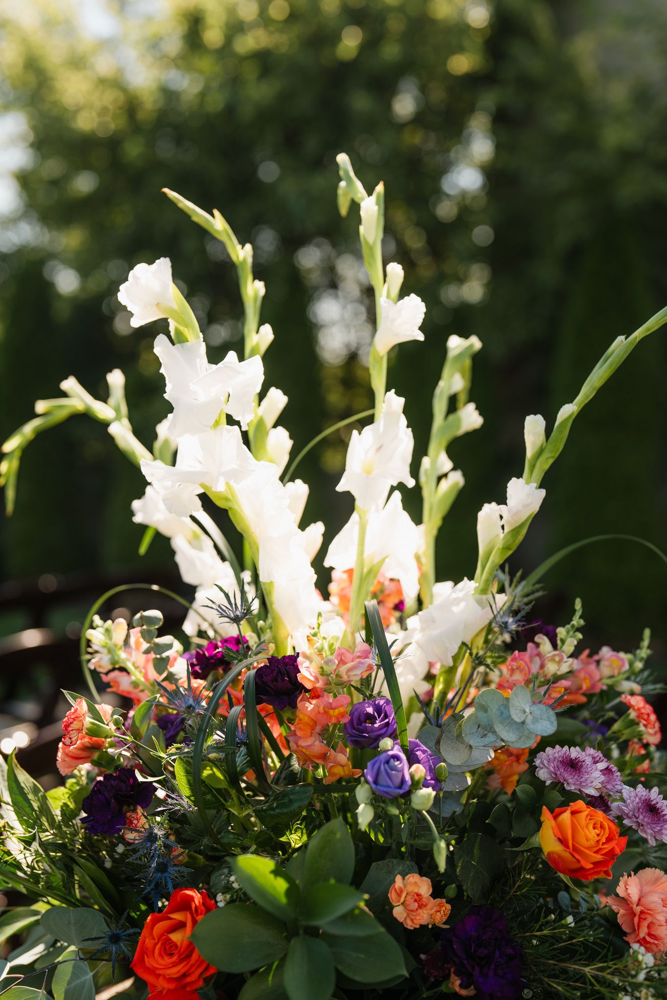 Vibrant floral arrangement featuring white gladiolus, orange roses, and purple flowers against a green bokeh background.