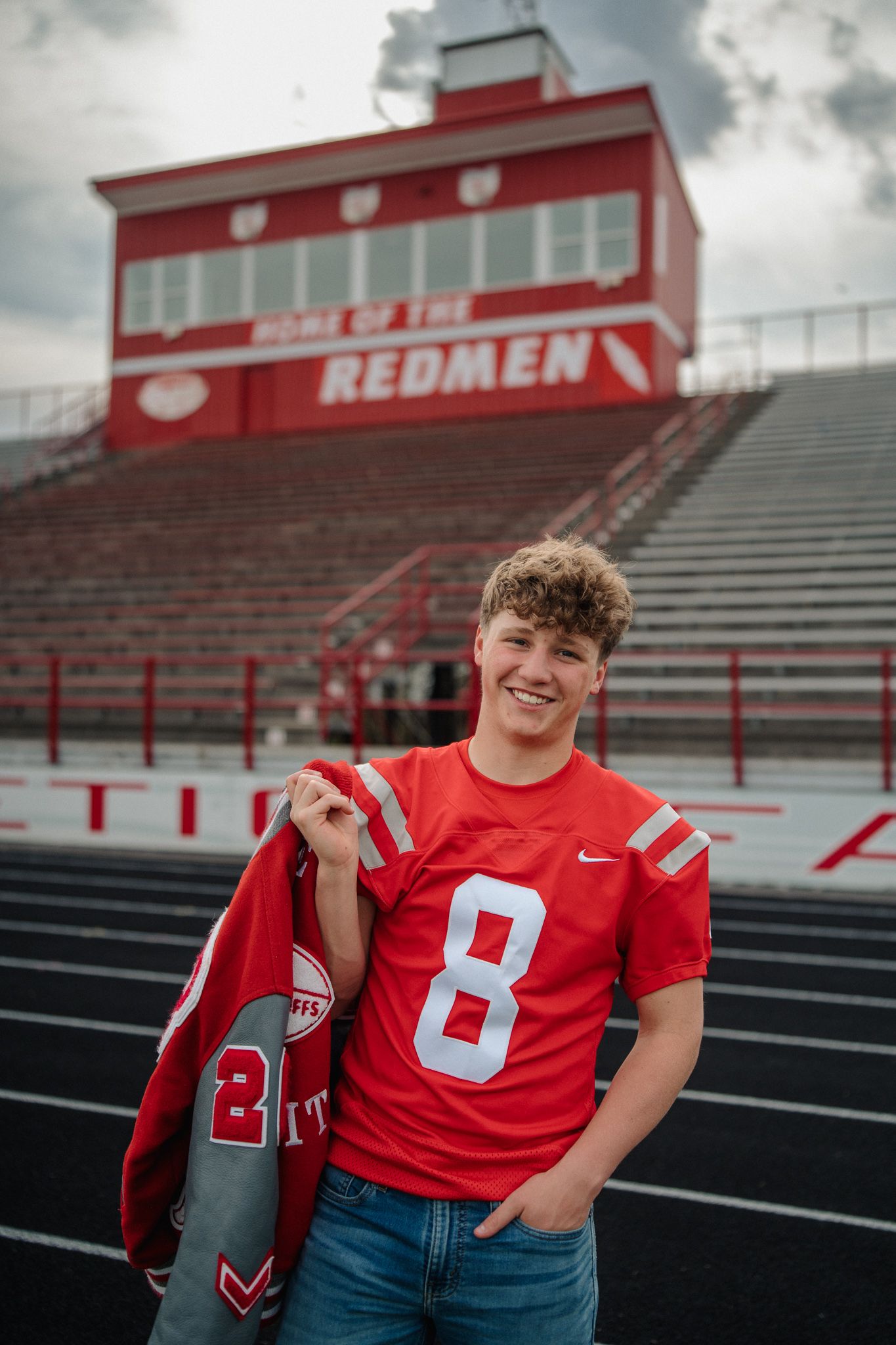Teen boy in red number 8 Nike jersey poses at Redmen football stadium bleachers for senior photos.