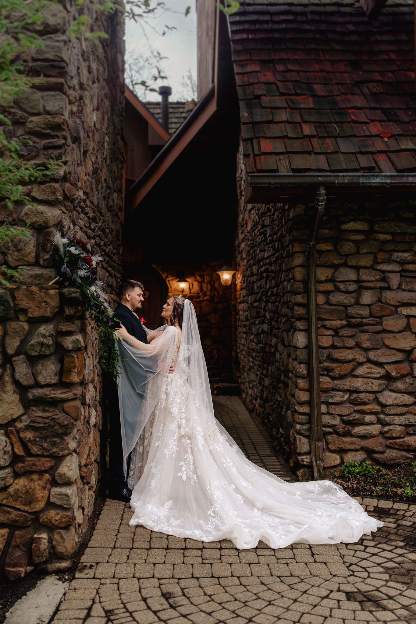 Bride and groom embrace in a romantic stone archway, her lace gown and veil trailing on cobblestone path.