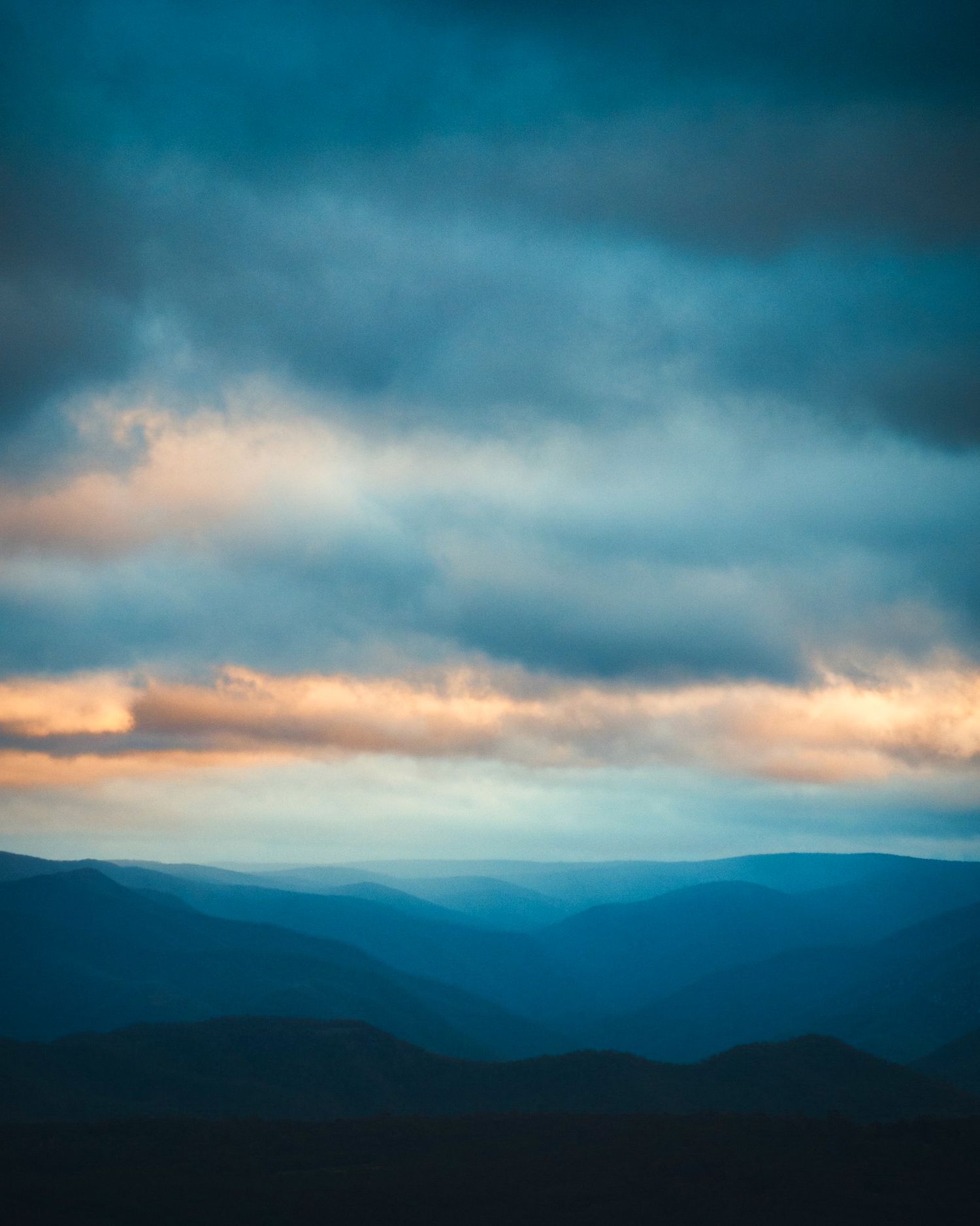 Dramatic cloudy sky over misty mountain ranges in moody blue tones at dusk.