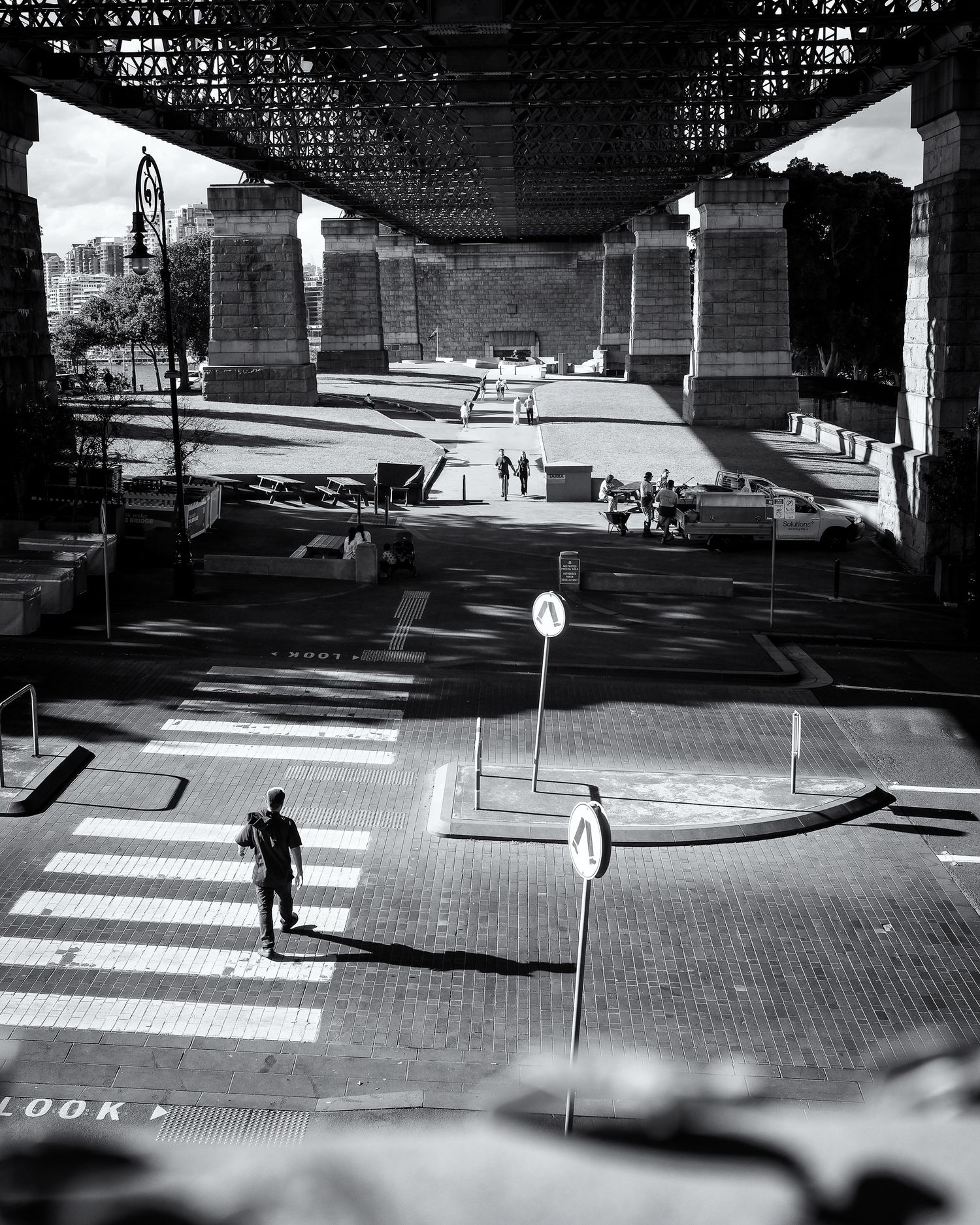 Black and white photo of a person walking under a large bridge near a crosswalk with road signs and urban architecture.