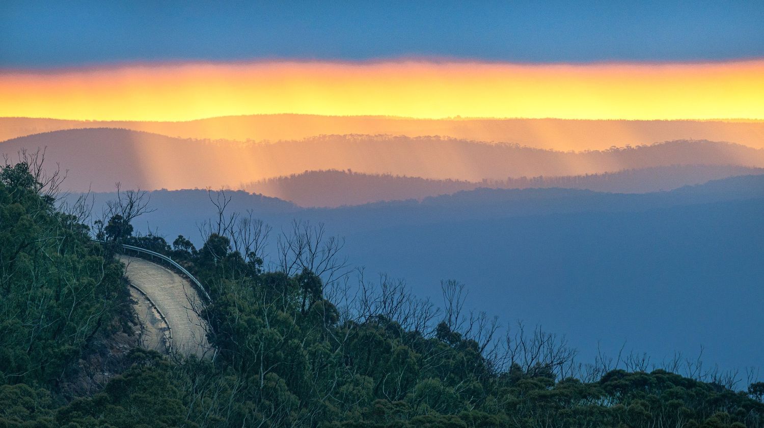 Stunning sunset over misty mountain layers with golden sky, green foreground shrubs, and a winding road in blue haze.