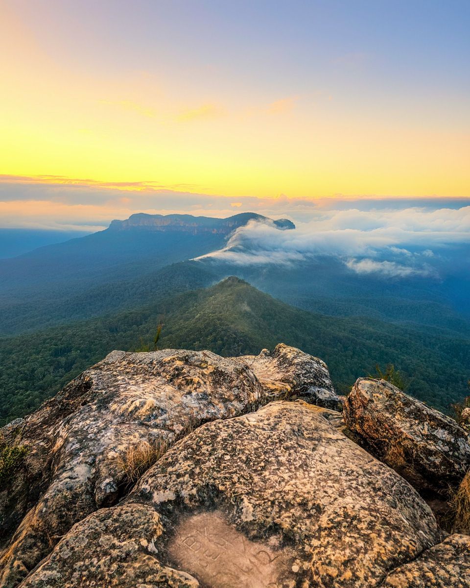 Rocky mountain summit overlooks lush green valleys with low clouds and a golden sunrise sky in the background.