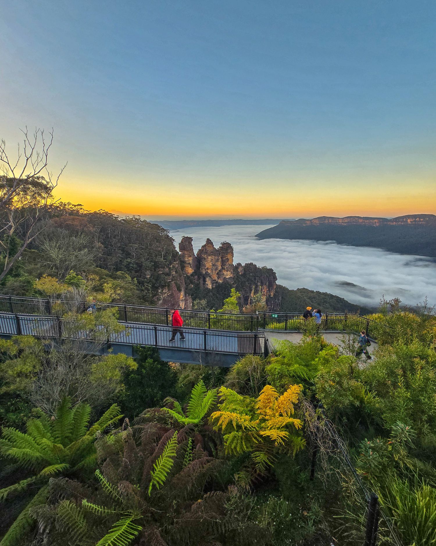 Tourists on a scenic walkway overlook the Three Sisters rock formation at sunrise in the Blue Mountains, Australia.