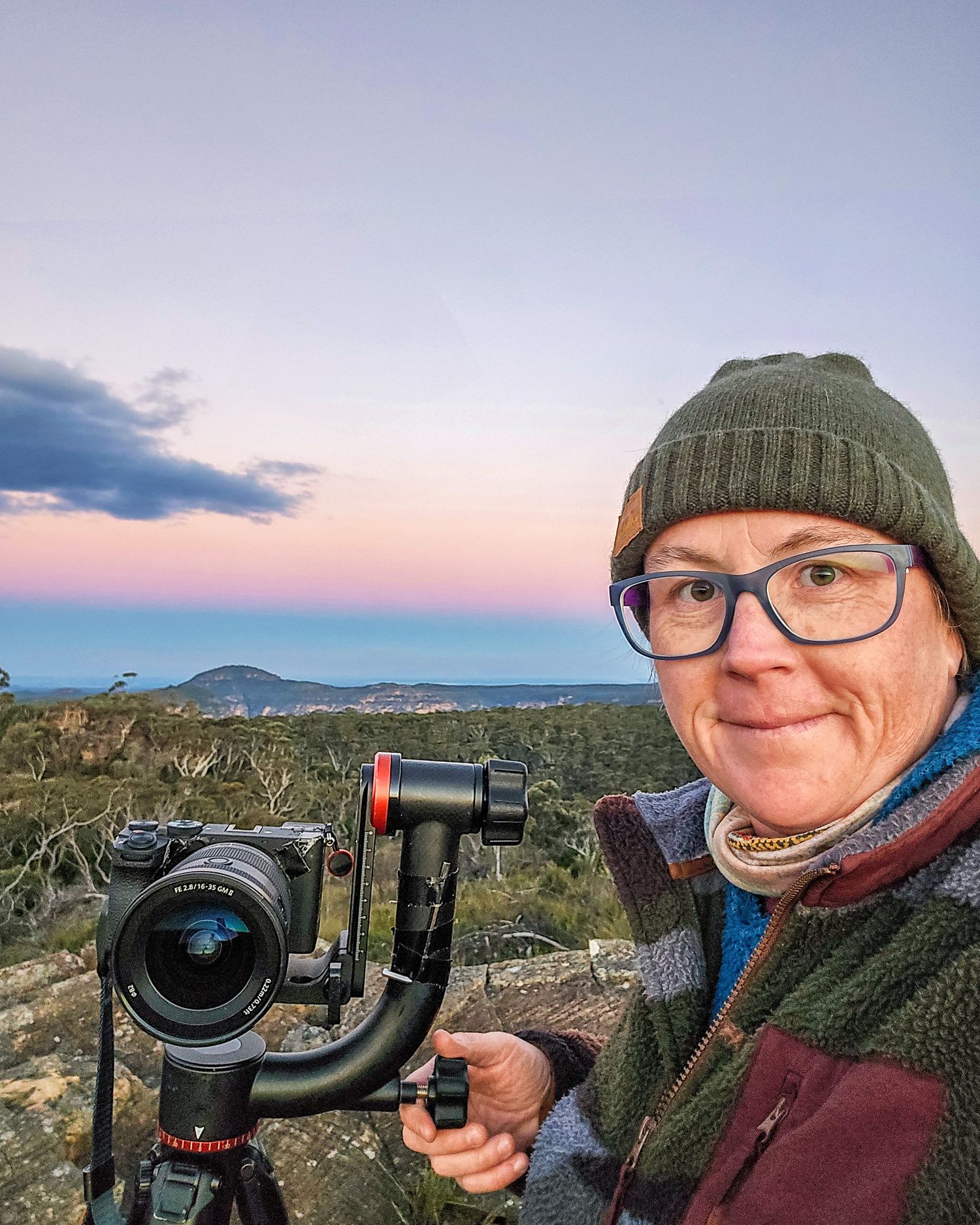 Person in green beanie and glasses holds camera gimbal outdoors at dusk with scenic coastal landscape behind.