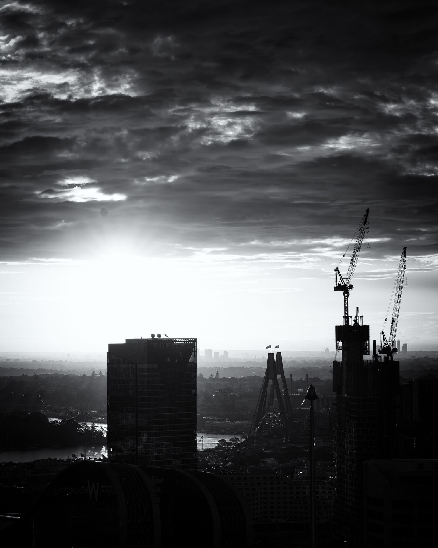 Black and white cityscape at sunset with construction cranes and dramatic cloudy sky silhouetting urban skyline.