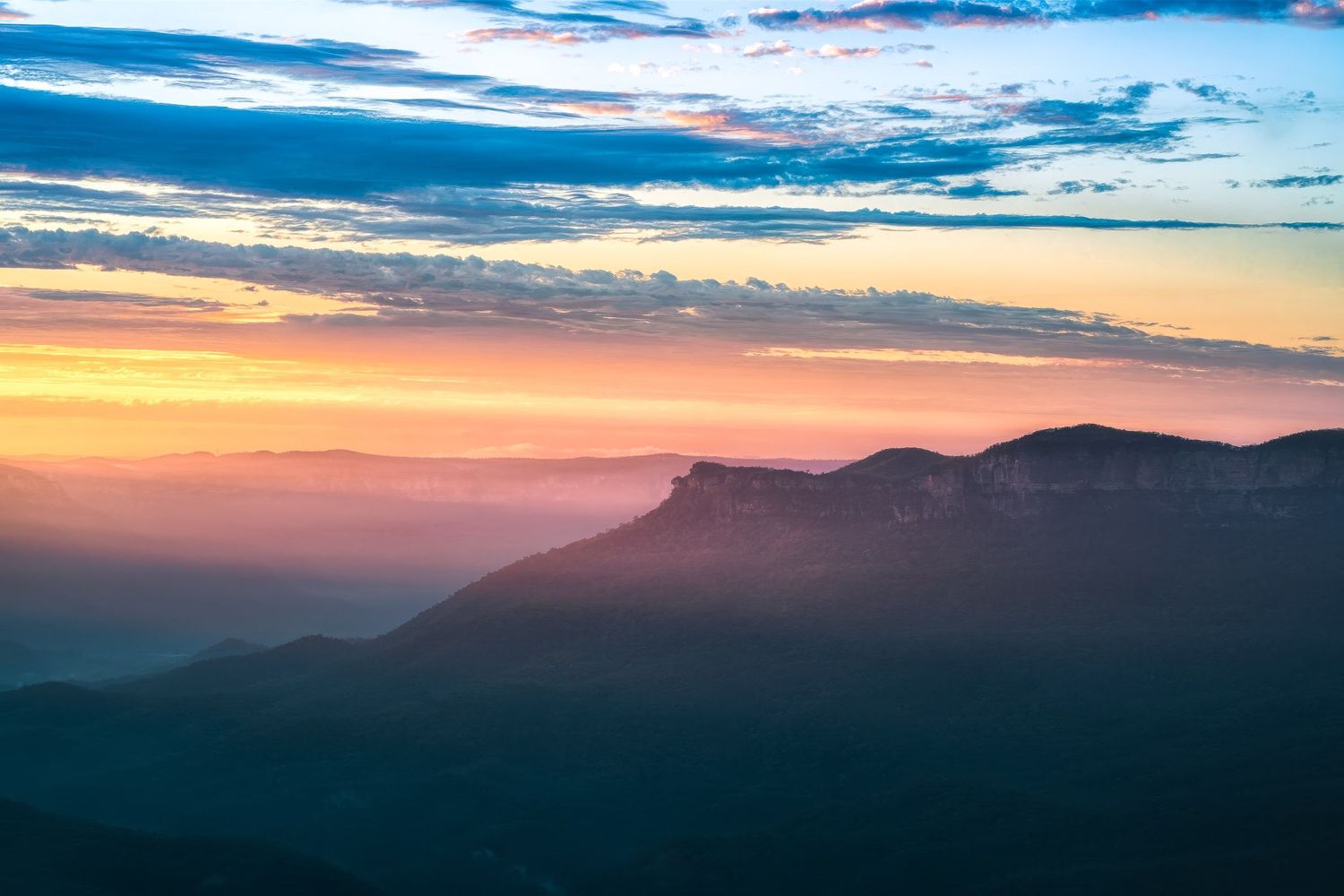 Stunning mountain sunset with vibrant orange and blue hues illuminating layered silhouettes of peaks under a dramatic cloudy sky.
